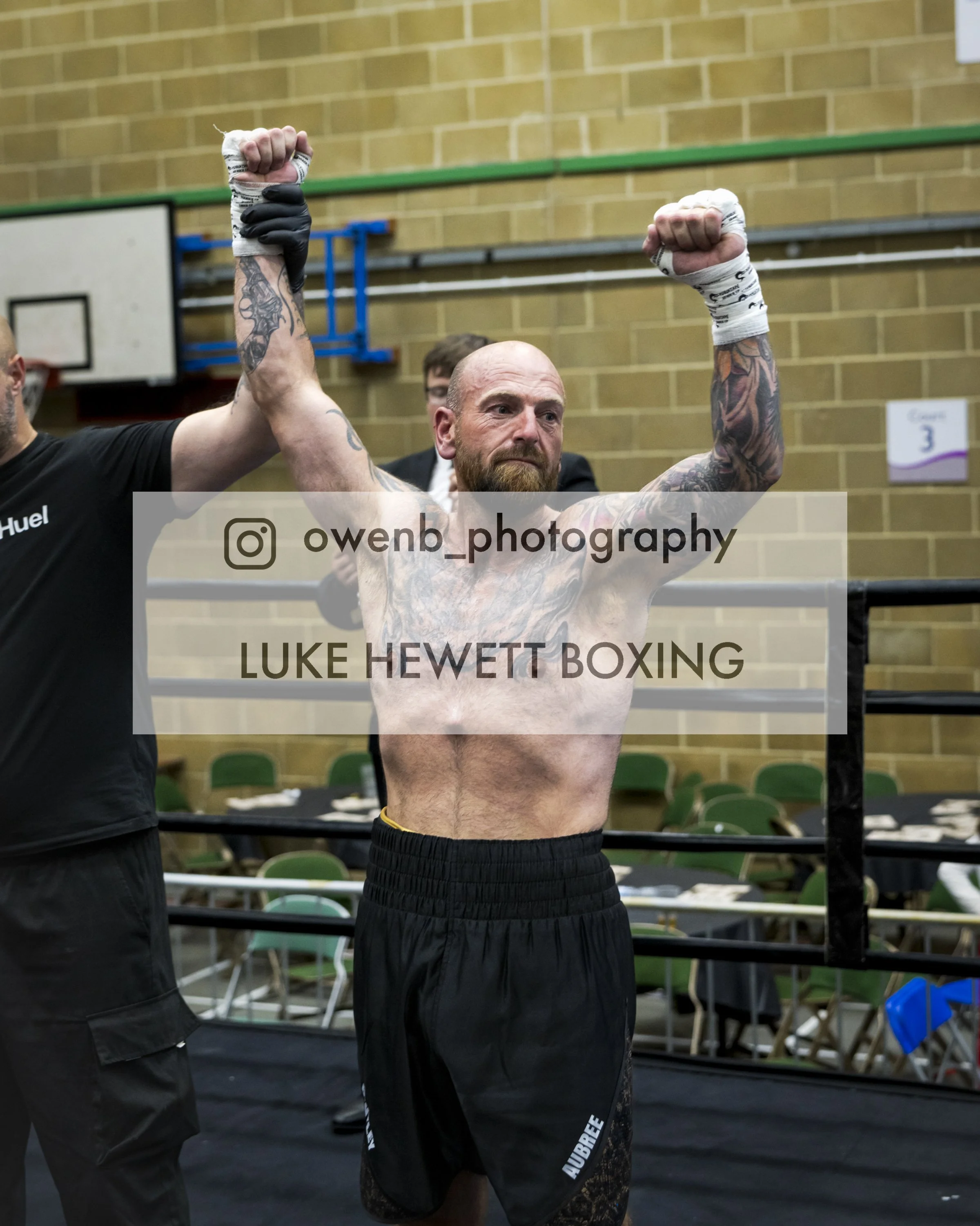 A shirtless male boxer with tattoos on his arms and chest, wearing black boxing shorts, stands in a boxing ring with his arms raised, holding his fists in the air as if celebrating or winning a fight. There is a referee or trainer partially visible t