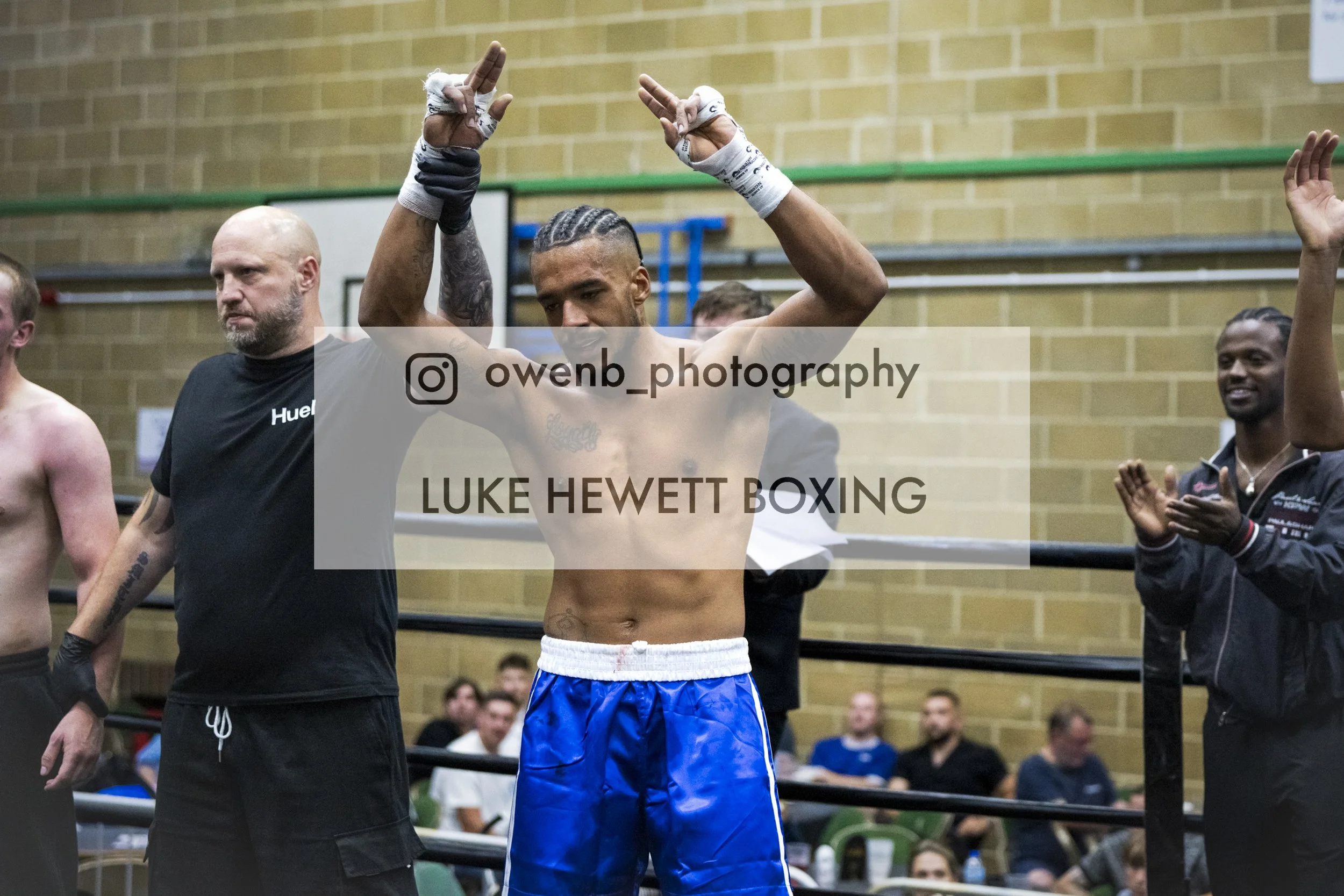 A shirtless male boxer with braided hair and tattoos, standing in a boxing ring with his hands raised, surrounded by spectators and officials.