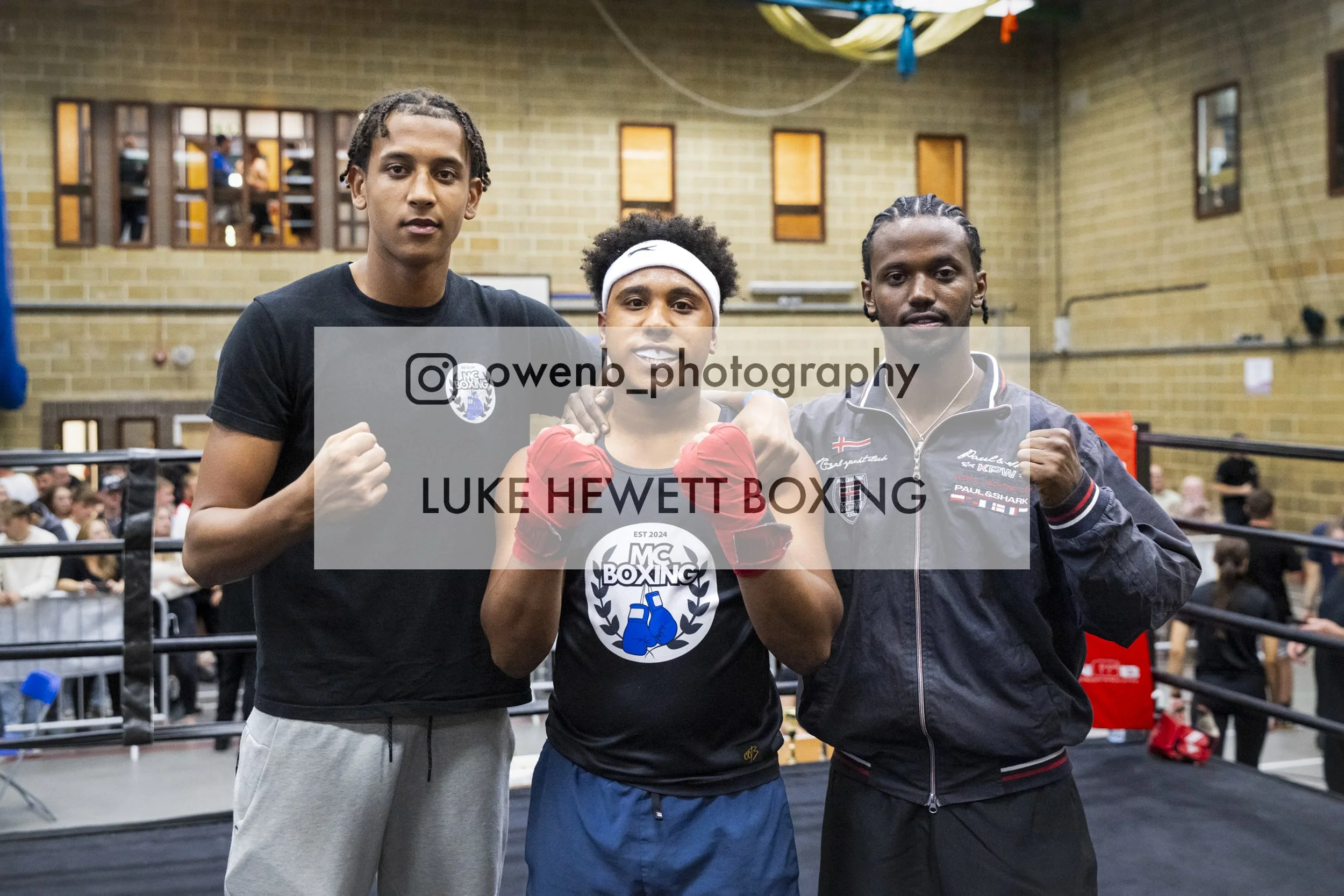Three men in boxing attire posing together in a boxing gym, with the central man making a fist and wearing boxing gloves with a logo, surrounded by a crowd and boxing ring.
