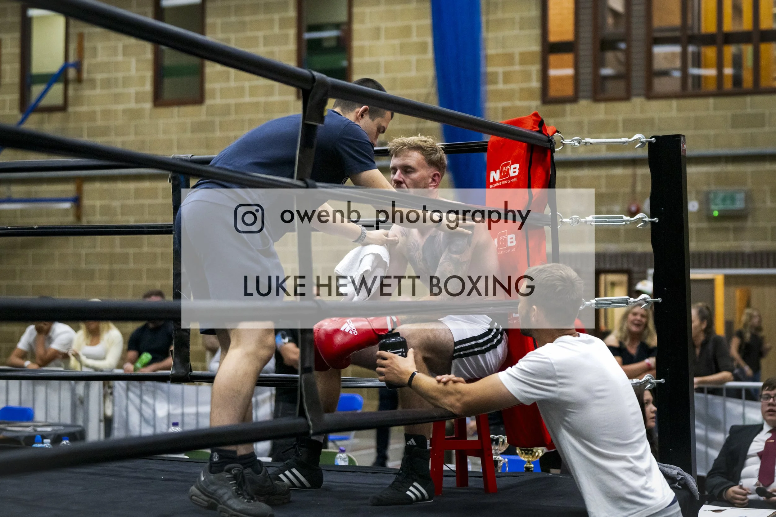 Boxer sitting on a stool in his corner during a match, receiving advice from his coach inside a boxing ring with an audience in the background.