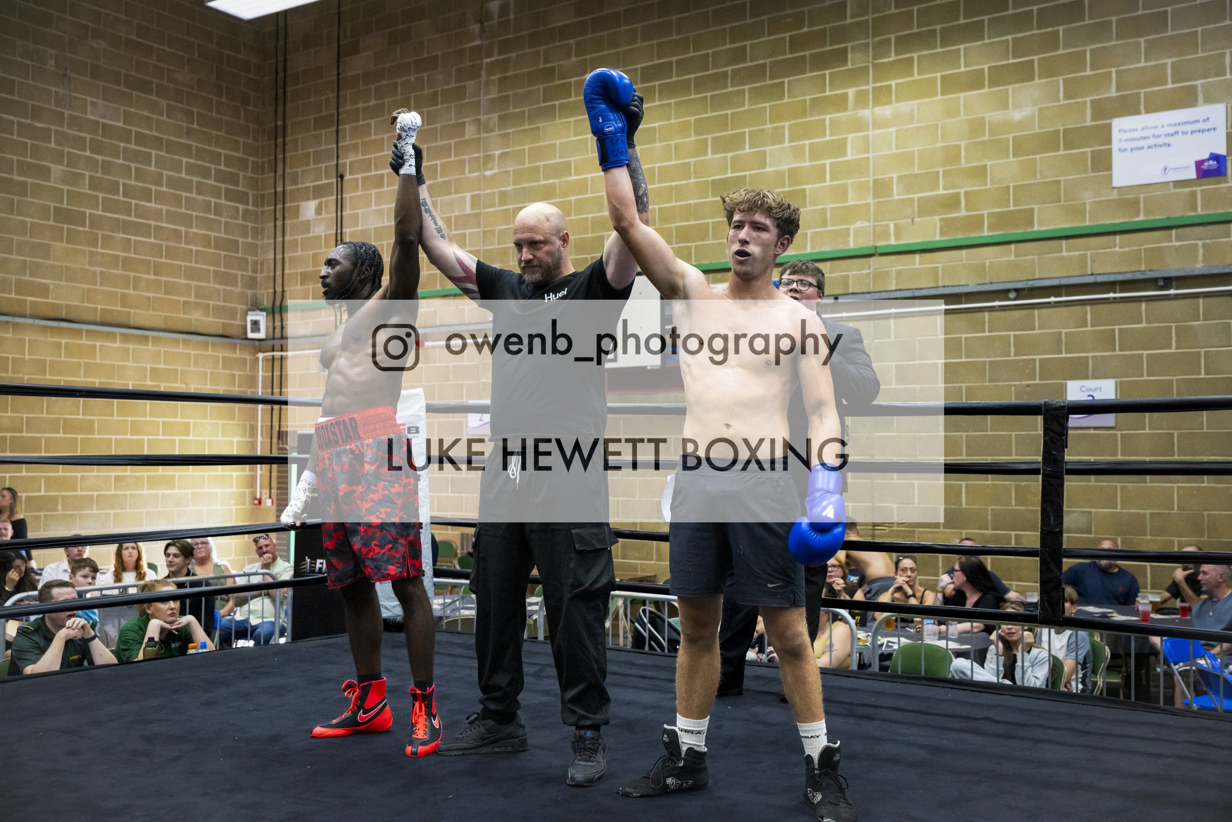 Boxers Luke Hewett and his opponent stand in the boxing ring after their match, with their arms raised by the referee. Audience members are seated around the ring, watching the event.