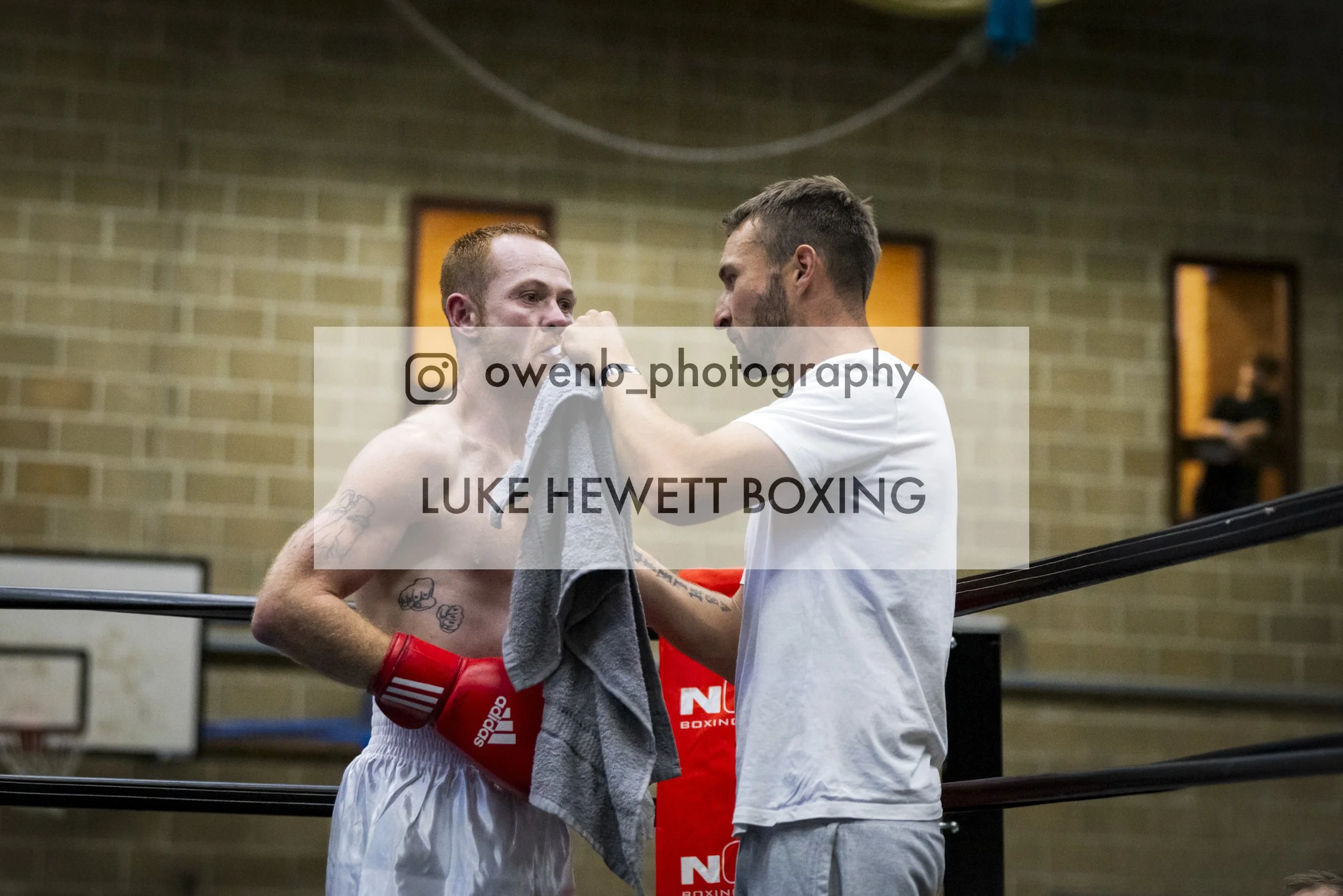 Two men, one shirtless boxer with tattoos and red gloves, and another man in a white t-shirt, are face-to-face in a boxing gym. The shirtless man is receiving encouragement or advice from the man in the white shirt. The background shows a brick wall 