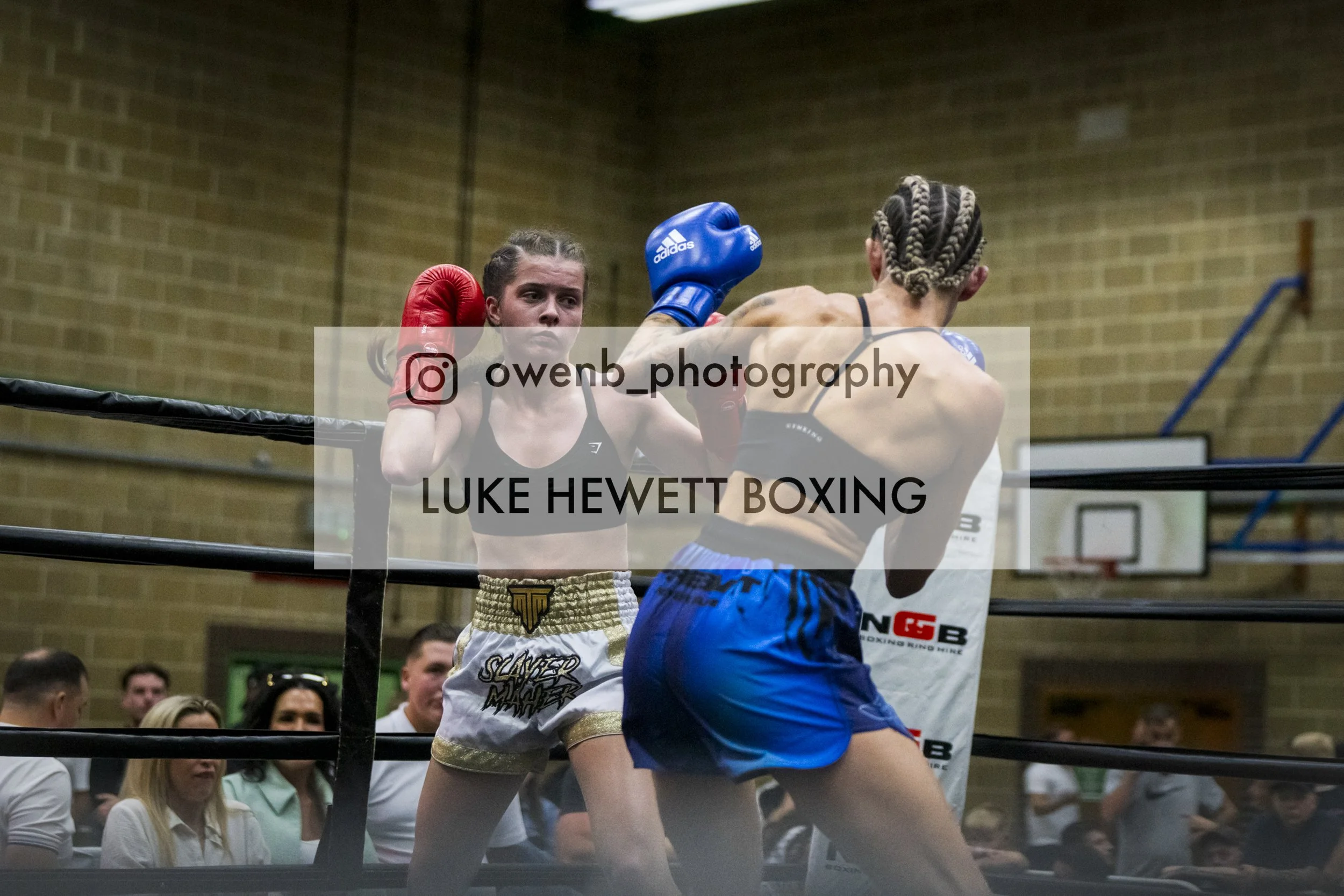 Two female boxers in a boxing match inside a gym, with one delivered a punch to the other, surrounded by an audience.