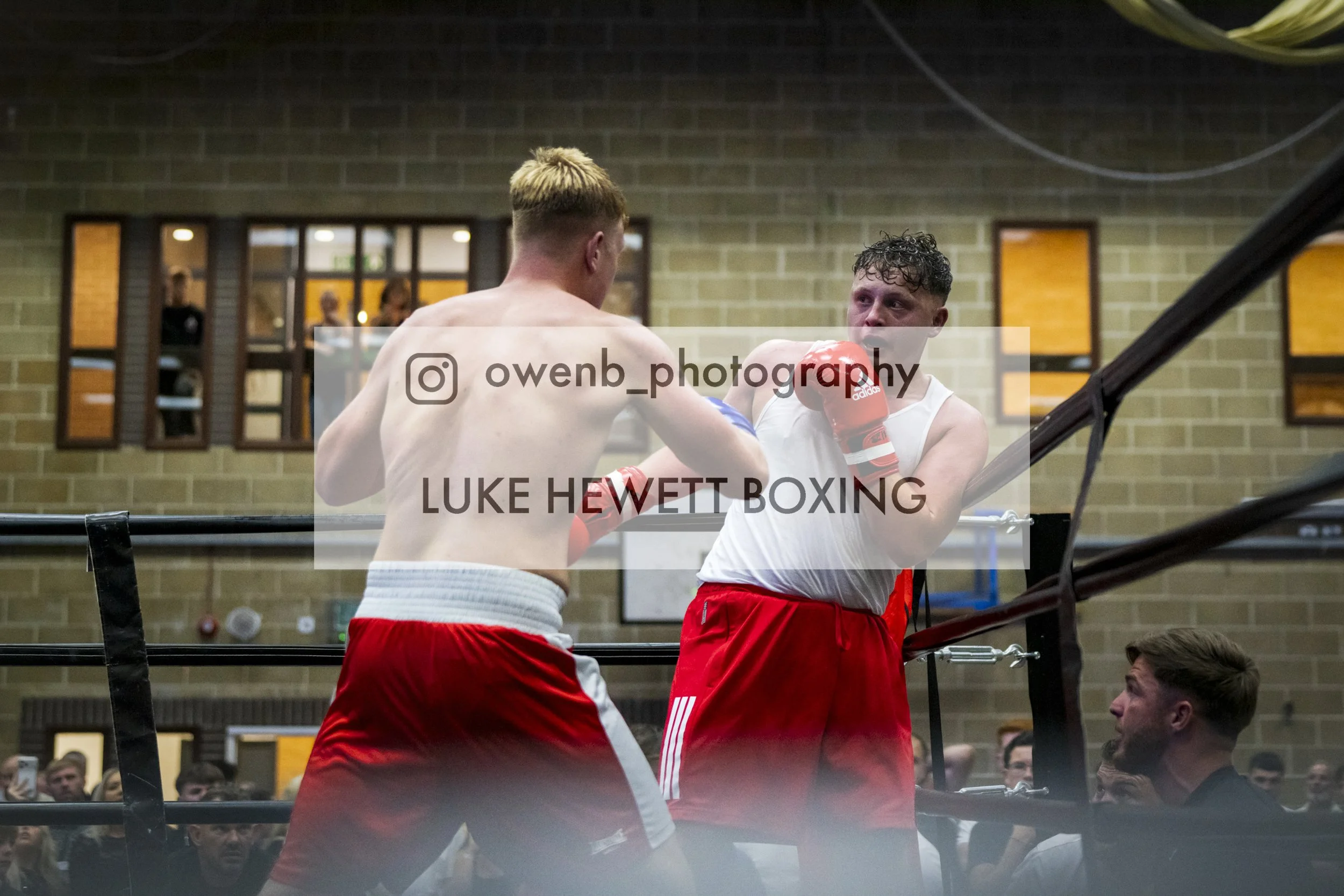 Two male boxers are sparring in a boxing ring indoors, with an audience watching, and their trainer standing nearby.