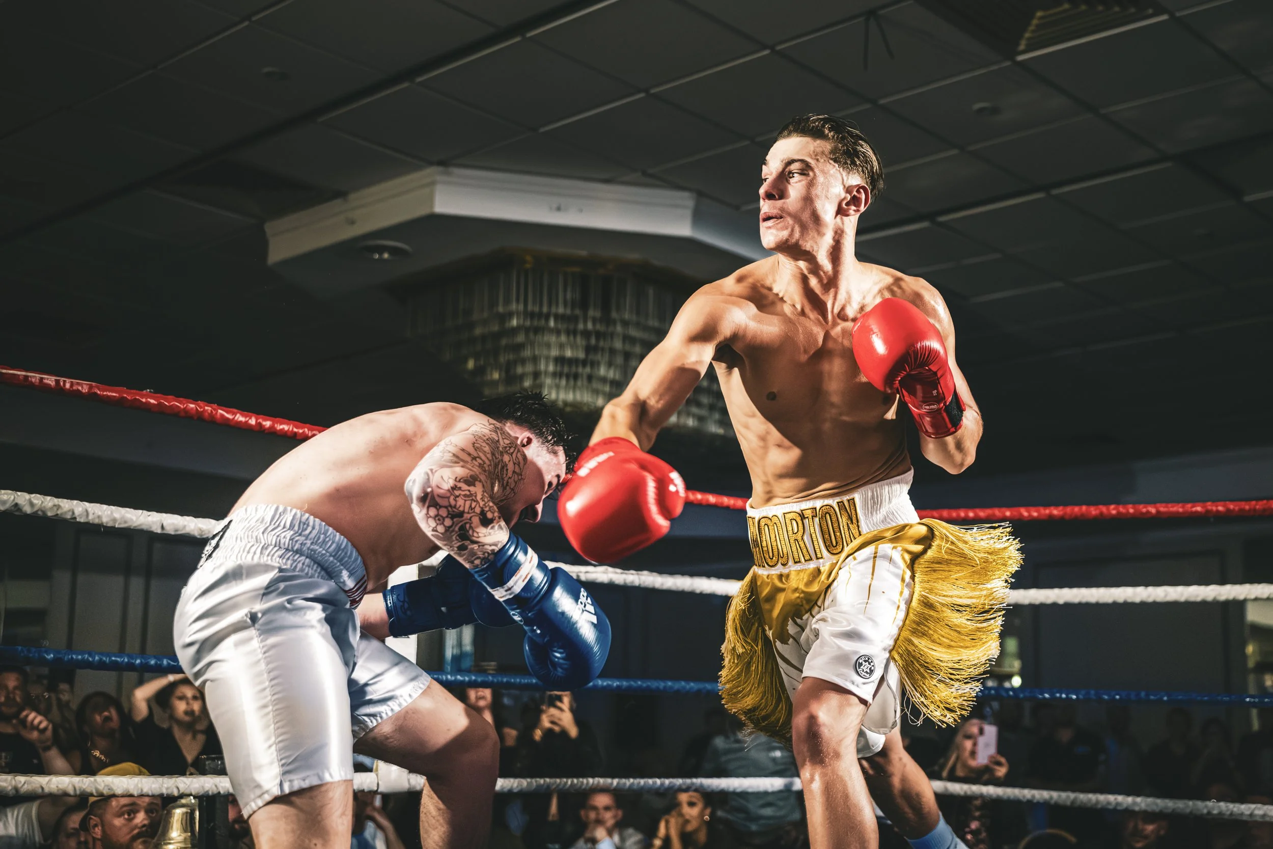 A boxer with a tattoo on his arm is getting punched in the midsection by a boxer wearing red gloves in a boxing ring, with spectators watching.