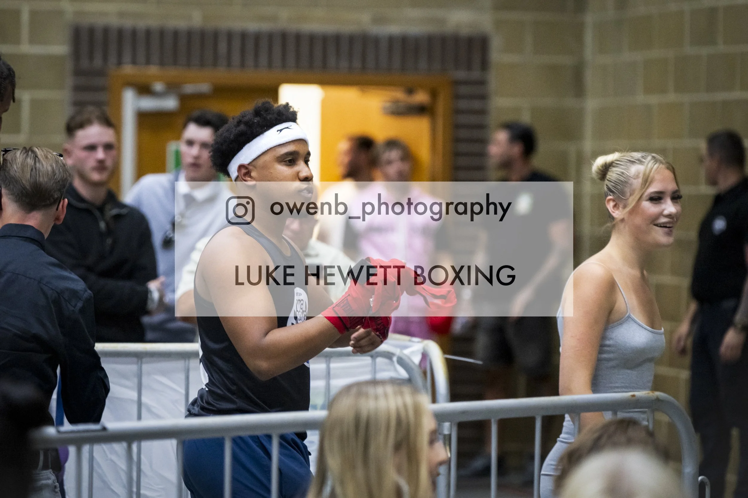 Young male boxer wearing red gloves and a headband, standing in a locker room, talking to a woman in a tank top, surrounded by other people.