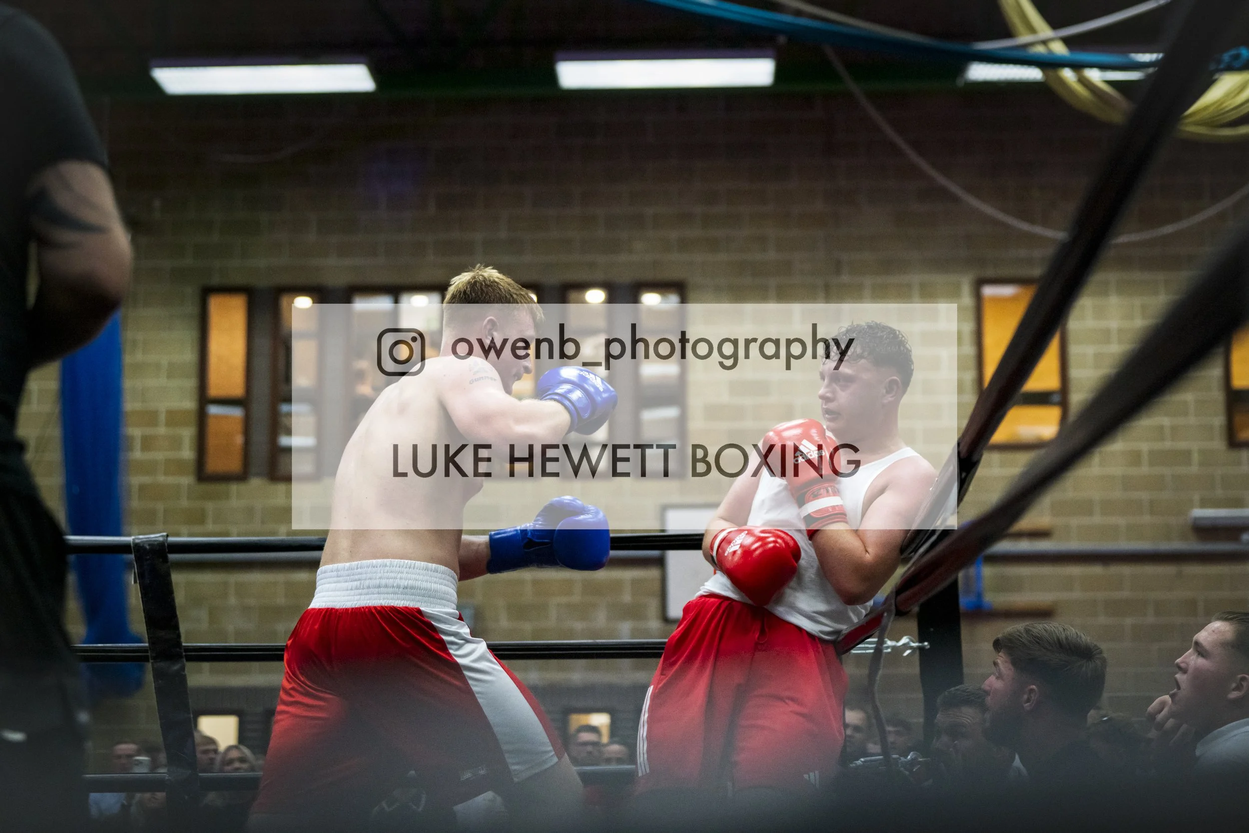 Two male boxers in a boxing ring, engaged in a bout, with one throwing a punch at the other, while spectators watch from below and around the ring.