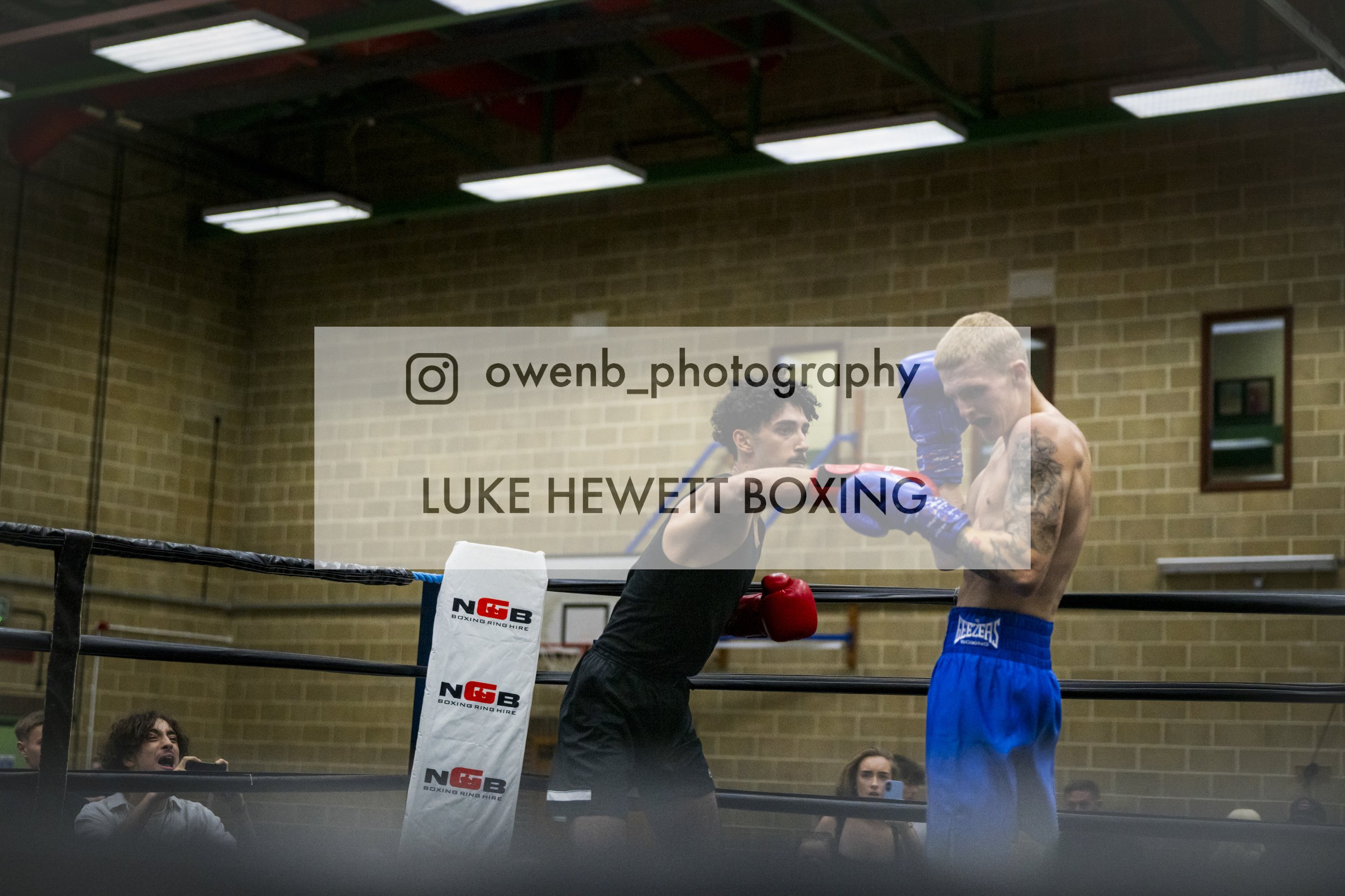 Two male boxers are sparring in a boxing ring, with one wearing red gloves and black shirt, the other in blue shorts and blue gloves. The scene is inside a gym with a brick wall background, and a few spectators can be seen watching and taking photos.