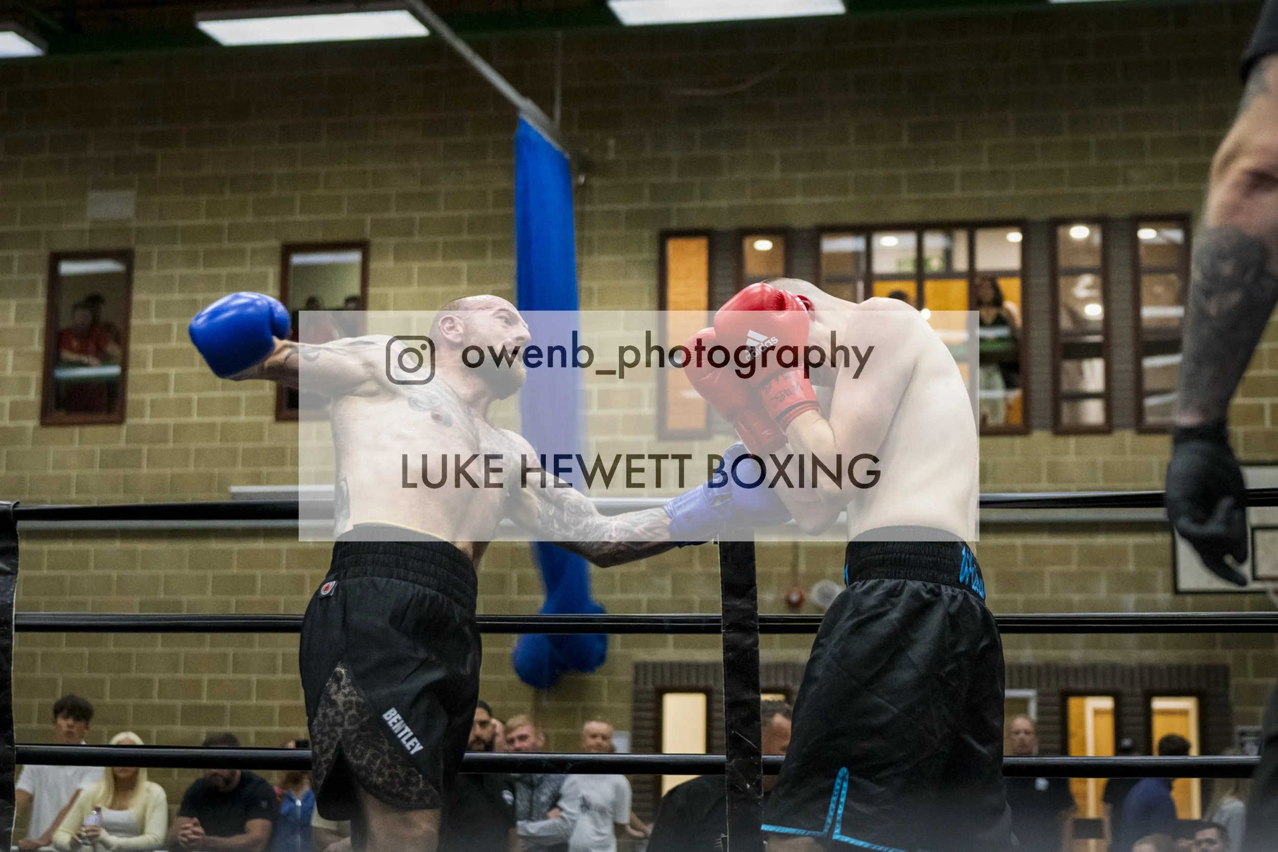 Two male boxers wearing boxing gloves and shorts are engaged in a match inside a boxing ring, with a referee observing. The background shows spectators and a brick wall with windows.