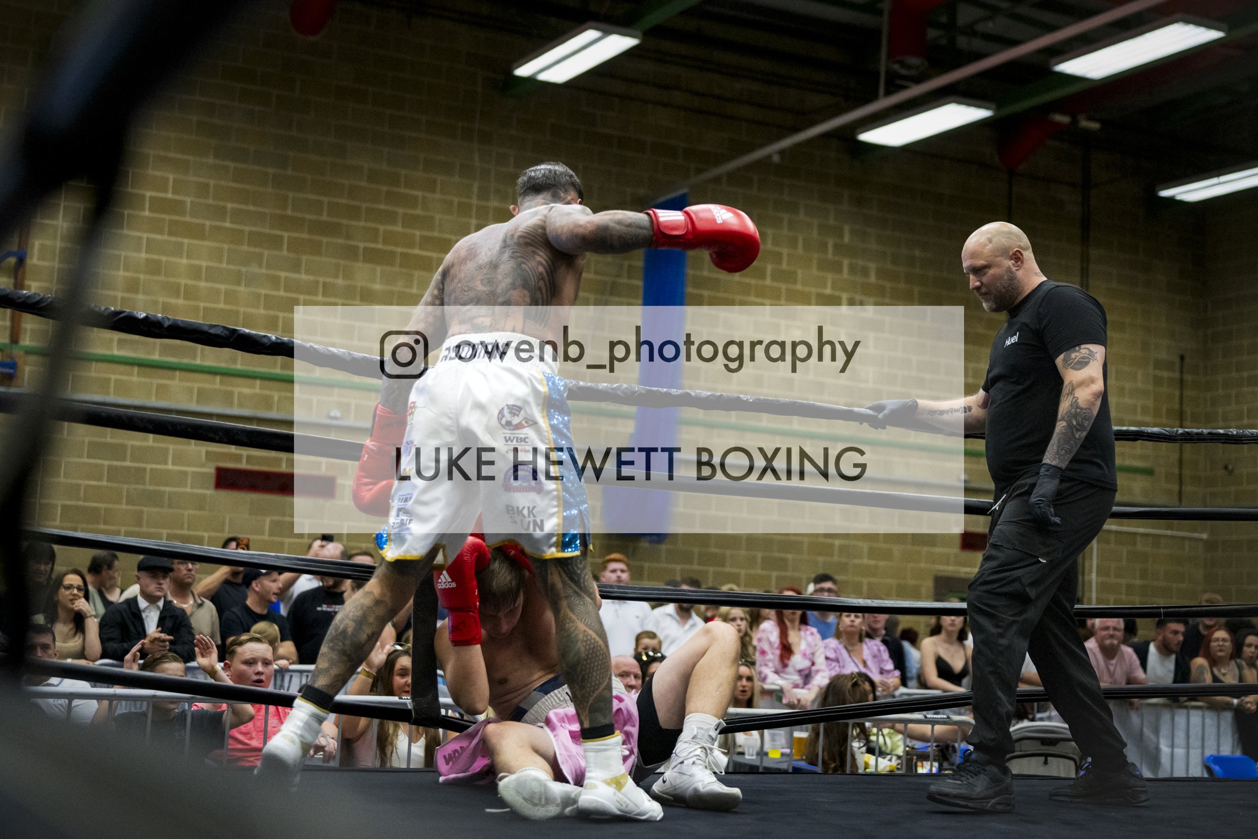 A boxing match with one fighter holding the other down on the ring floor, while the referee looks on. The crowd watches in the background inside a gym or sports hall.