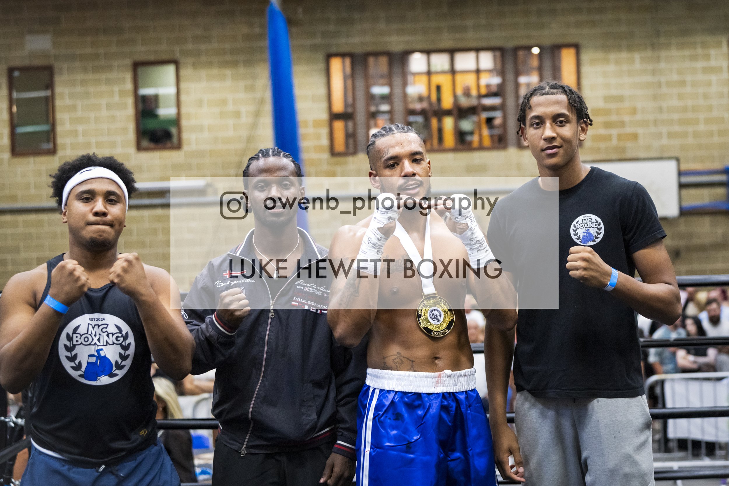 Four male boxers standing in a boxing gym, posing for a photo. The boxer second from the right is wearing a gold medal around his neck. The others have their fists raised in a fighting stance. The gym has a yellow brick wall and windows in the backgr