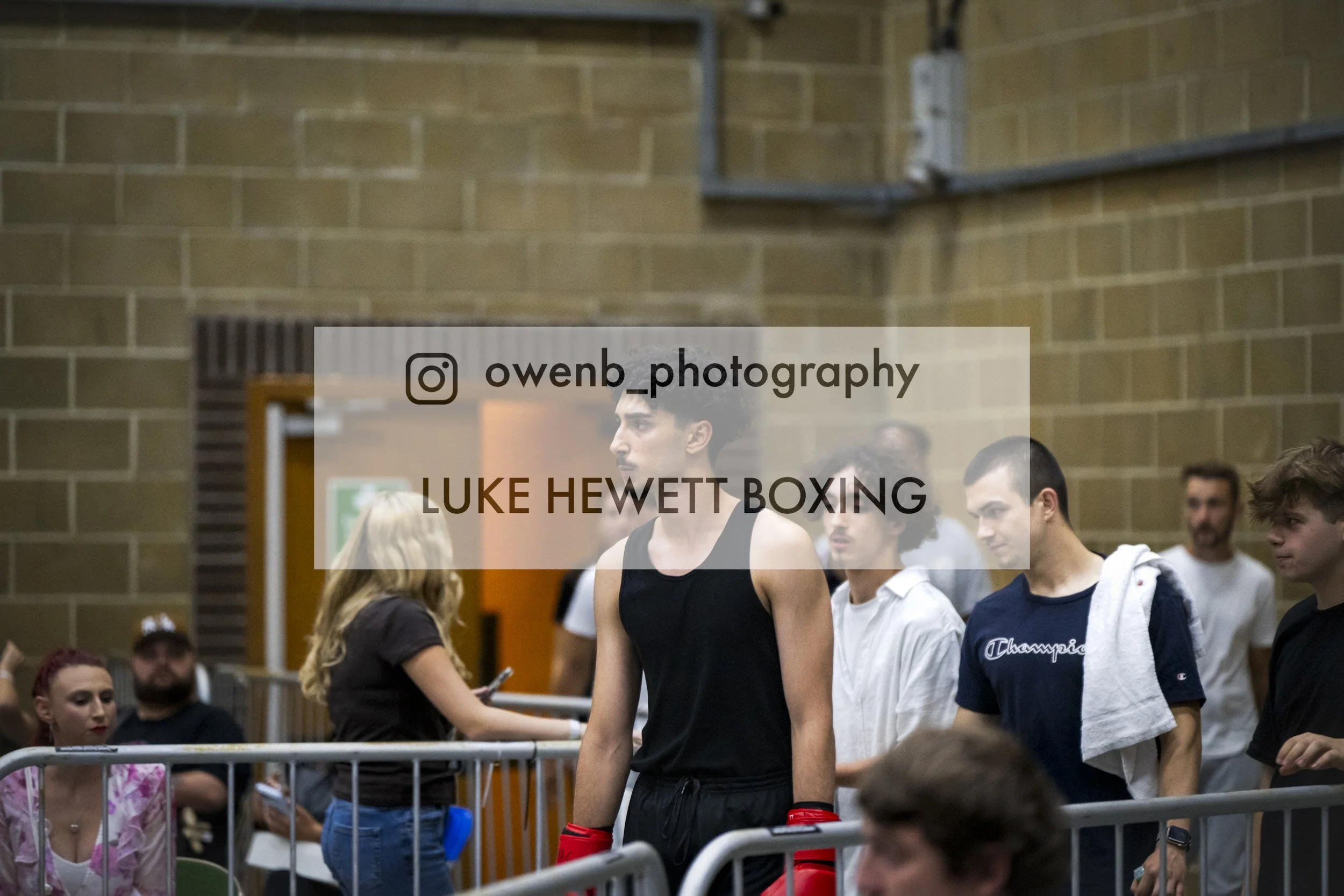 A young male boxer with dark hair and a black tank top standing in a gym, surrounded by people, possibly at a boxing event or training session.