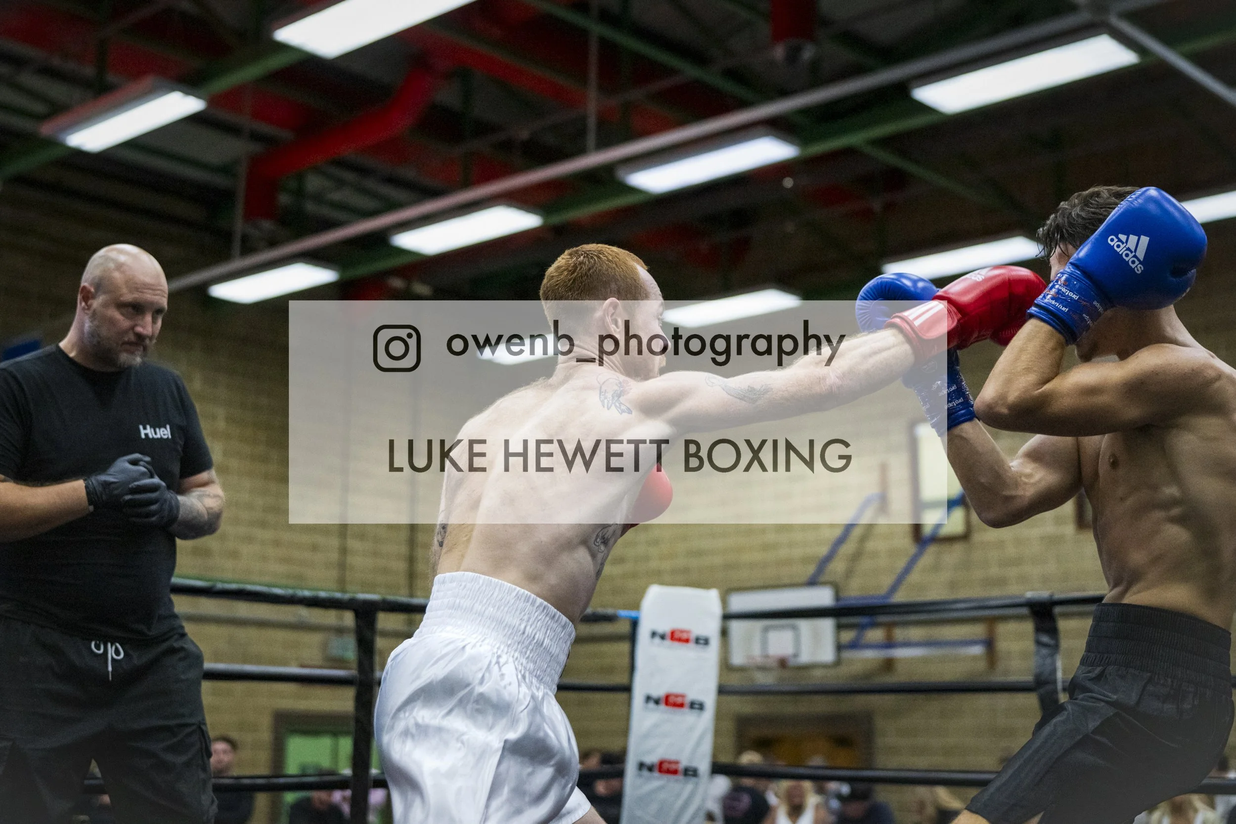 Two shirtless male boxers, wearing gloves, are sparring in a boxing ring. One is throwing a punch while the other is covering their face. An official or coach, wearing black, observes nearby, with a wooden gym wall and overhead lights in the backgrou