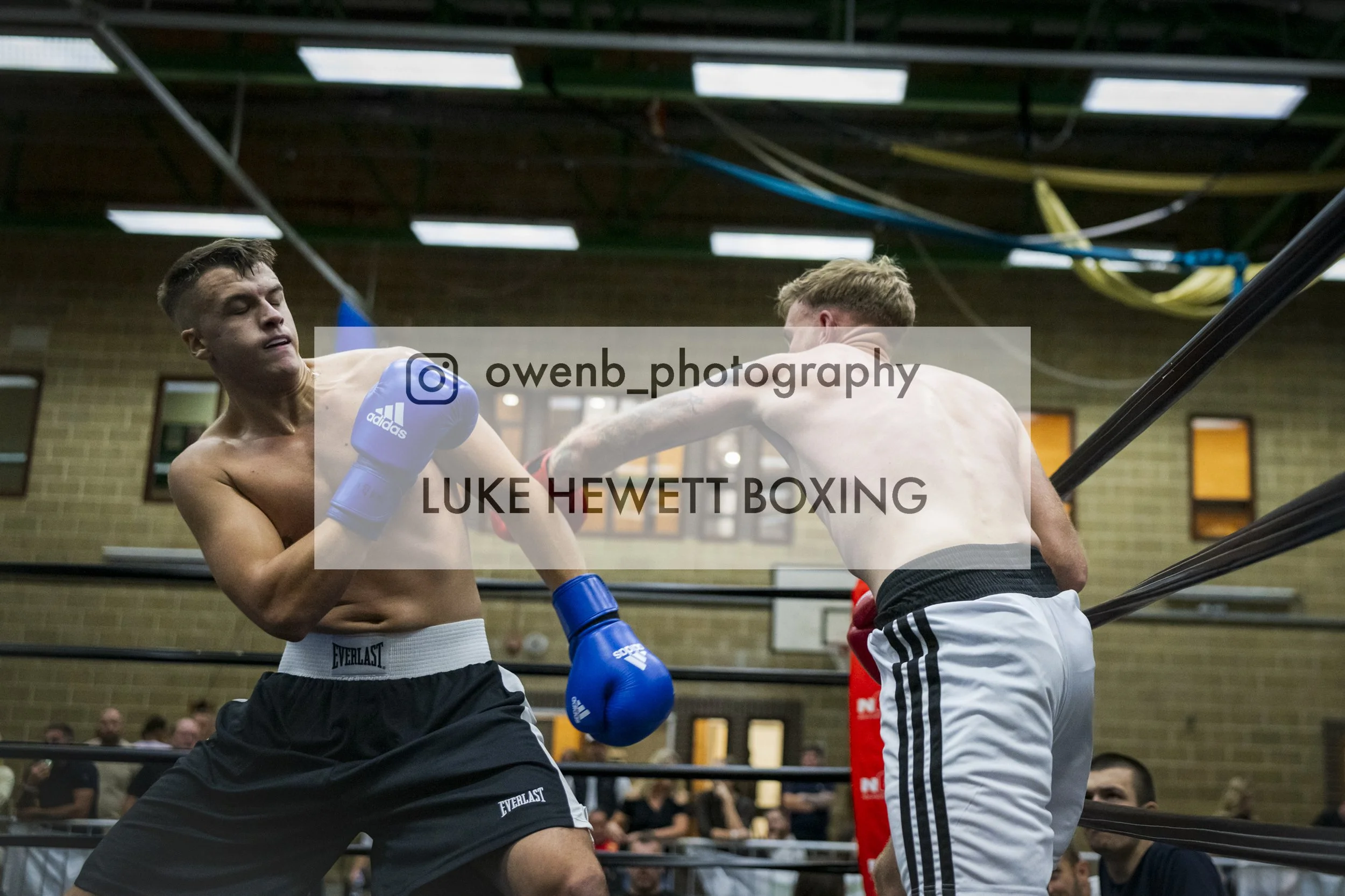 Two shirtless male boxers fighting in a boxing ring, with one delivering a punch to the other. Eyewear and gloves are visible, and a crowd watches in the background.