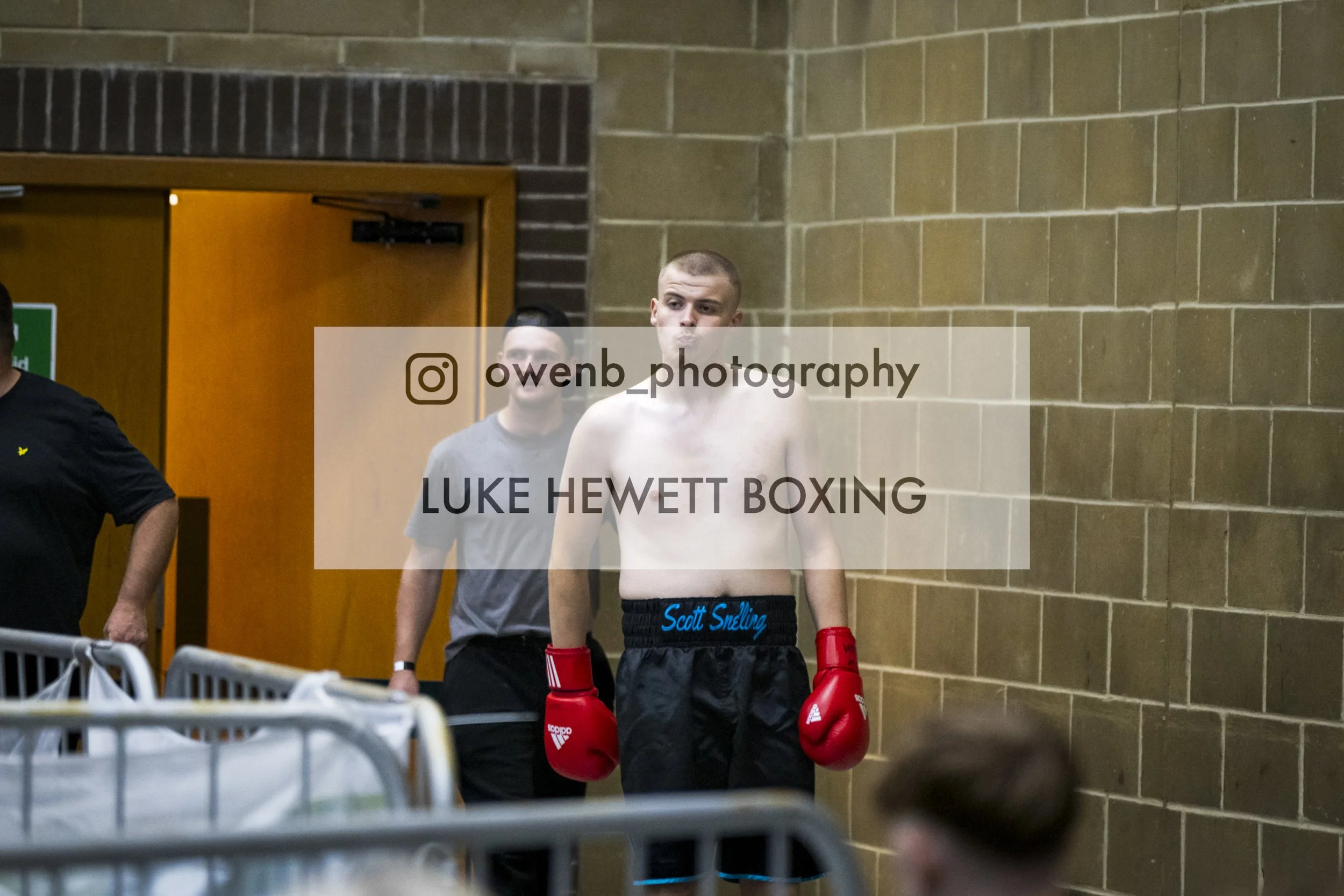 A young male boxer with boxing gloves and shorts, standing in a corridor with a brick wall, accompanied by two men, one of whom is shirtless.