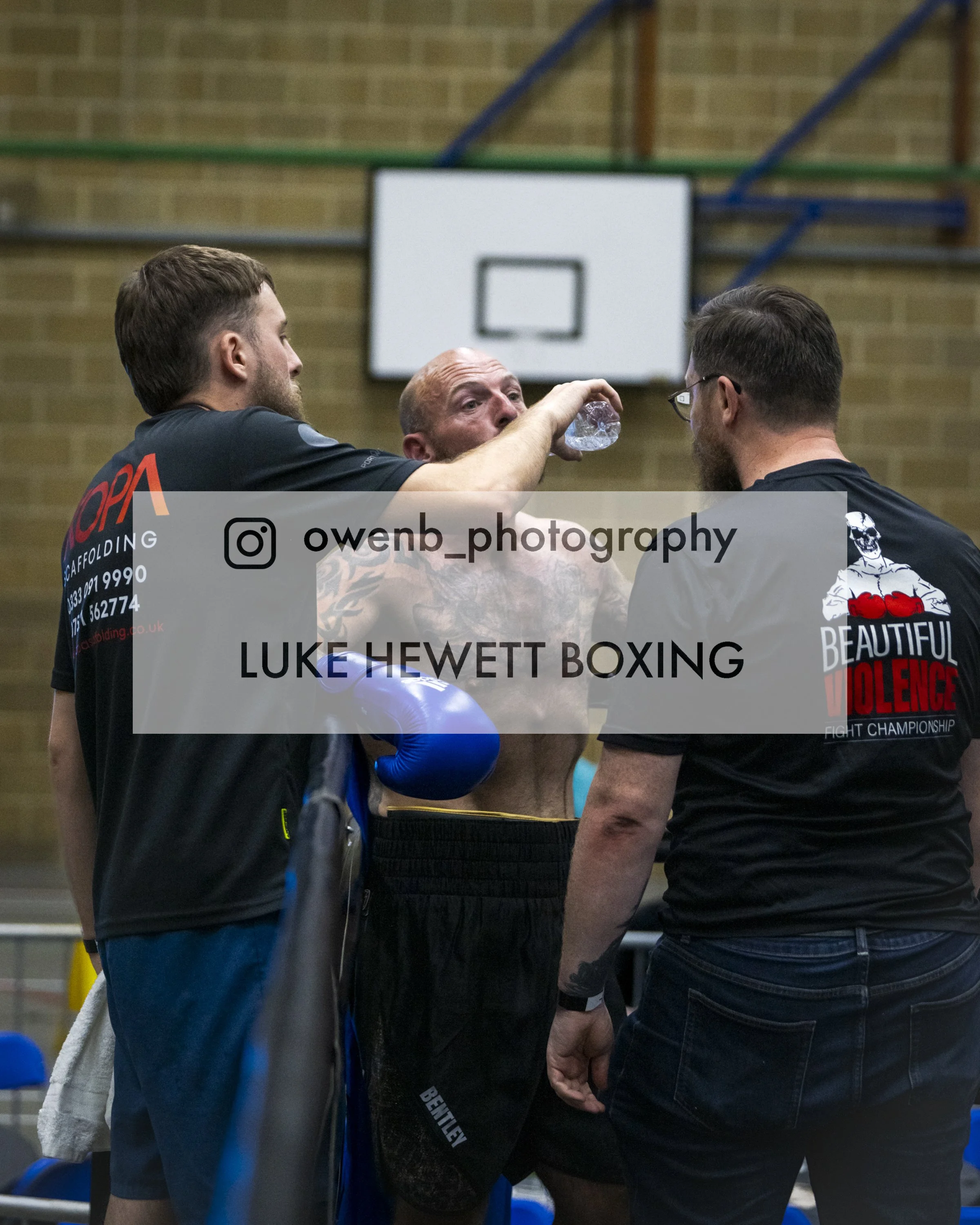 Three men in a gym or sports hall, standing in a close circle with one of them drinking water from a bottle, with a basketball hoop in the background.