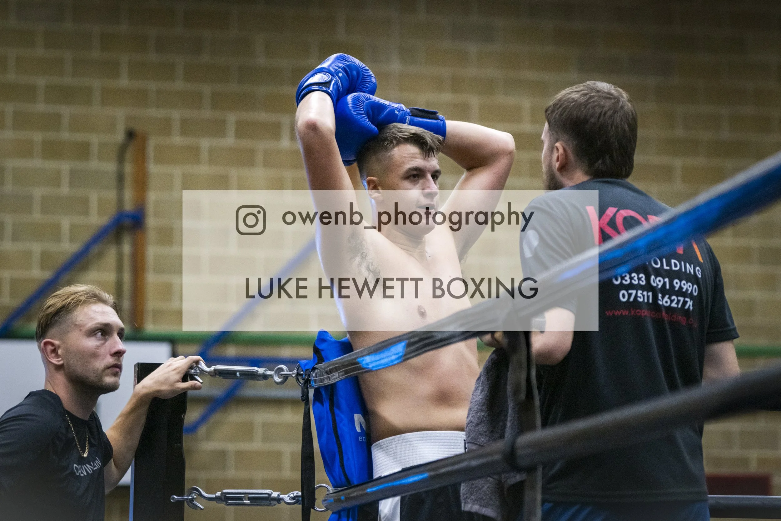 A boxer with a muscular build is being assisted by a trainer in a gym. The boxer is shirtless, wearing blue boxing gloves and shorts, and has his arms raised above his head. The trainer, with a beard and wearing a dark T-shirt, is holding the boxer's