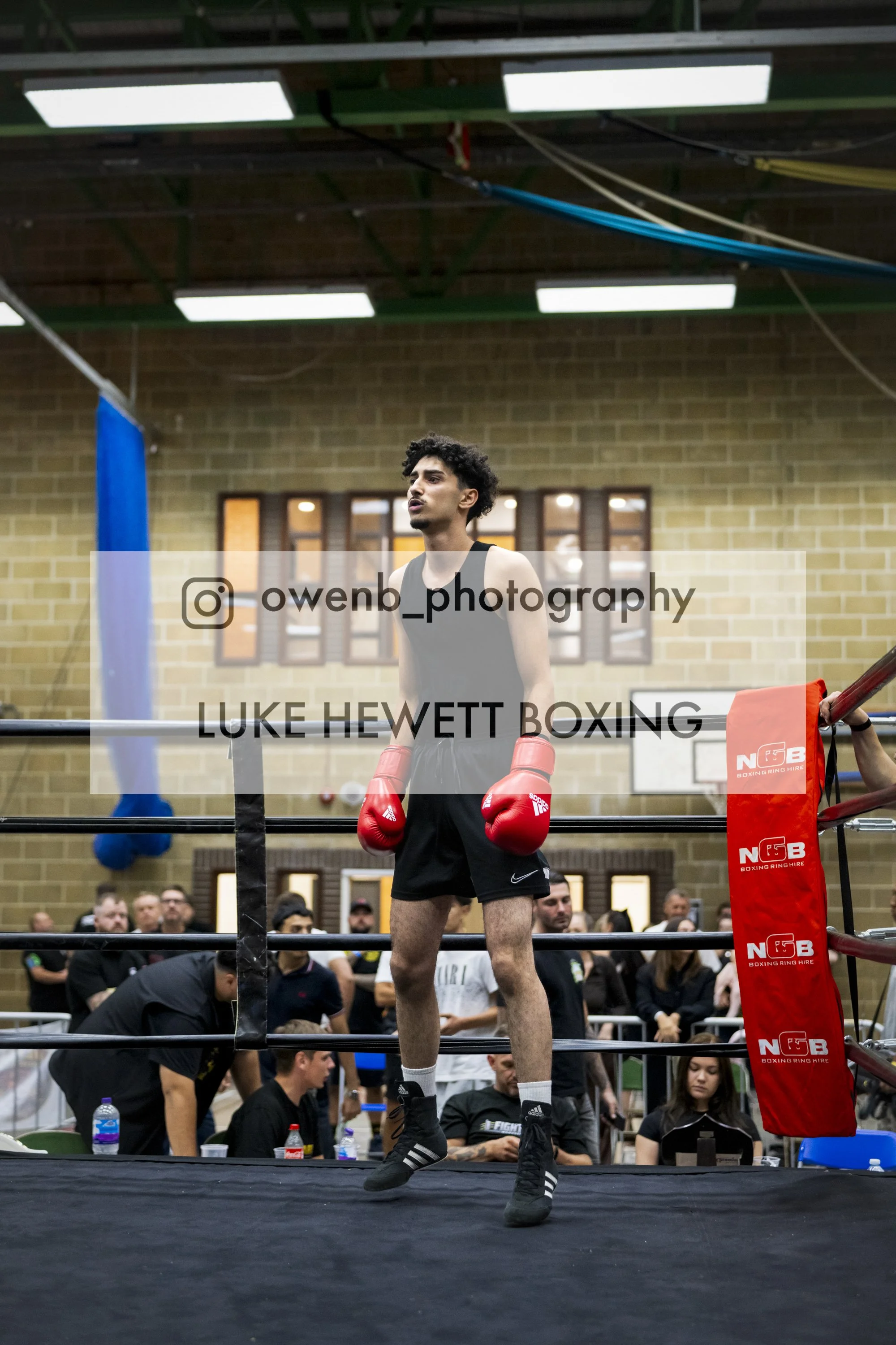 A male boxer stands in a boxing ring wearing red gloves and black shorts, with an attentive expression. Spectators are visible in the background at an indoor event.