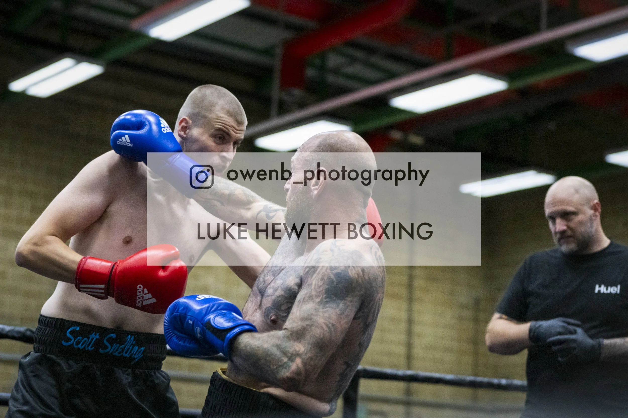Two male boxers, one with blue gloves and the other with red gloves, sparring in a gym, with one delivering a punch and the other blocking, while a coach observes in the background.