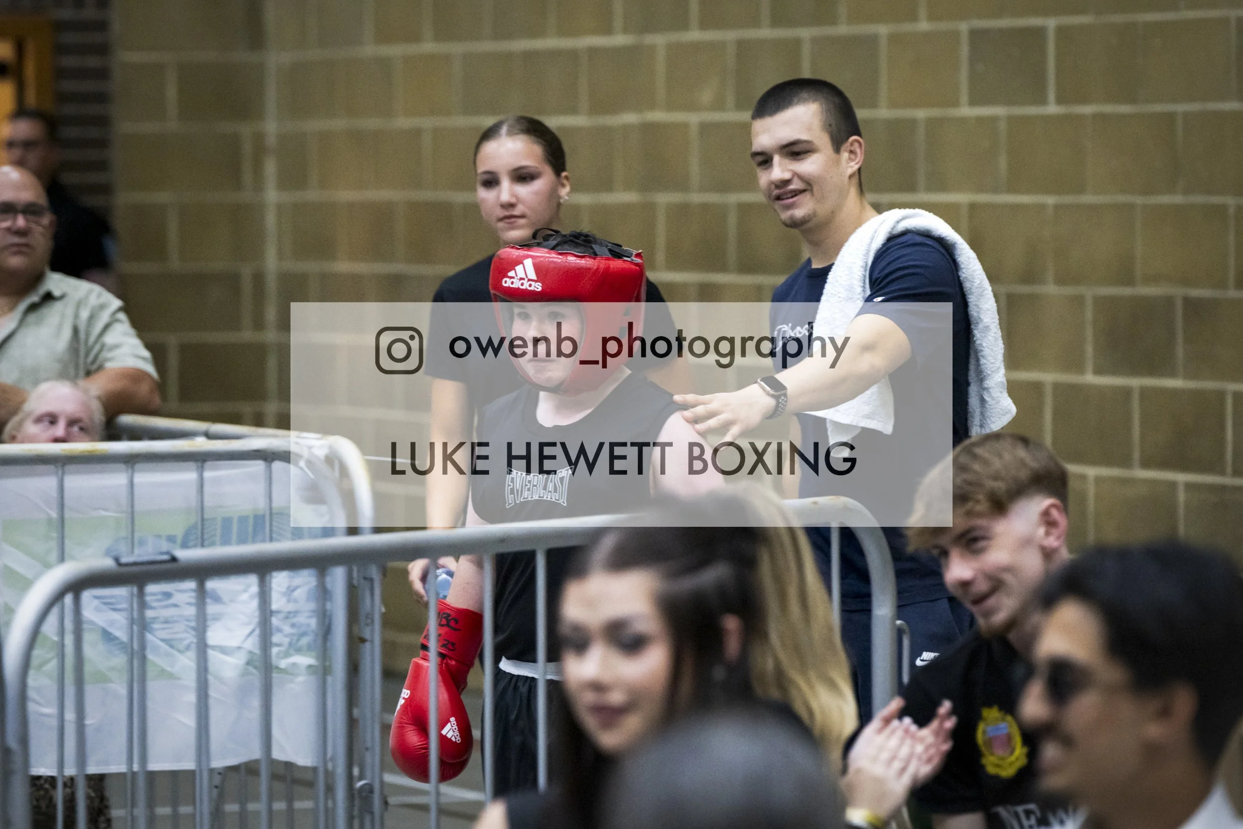 Inside a gym or boxing arena, a group of people are gathered. A young boxer girl dressed in black gear with red boxing gloves and headgear is in the center, accompanied by a woman, possibly a coach or trainer. The man on the right is smiling, wearing