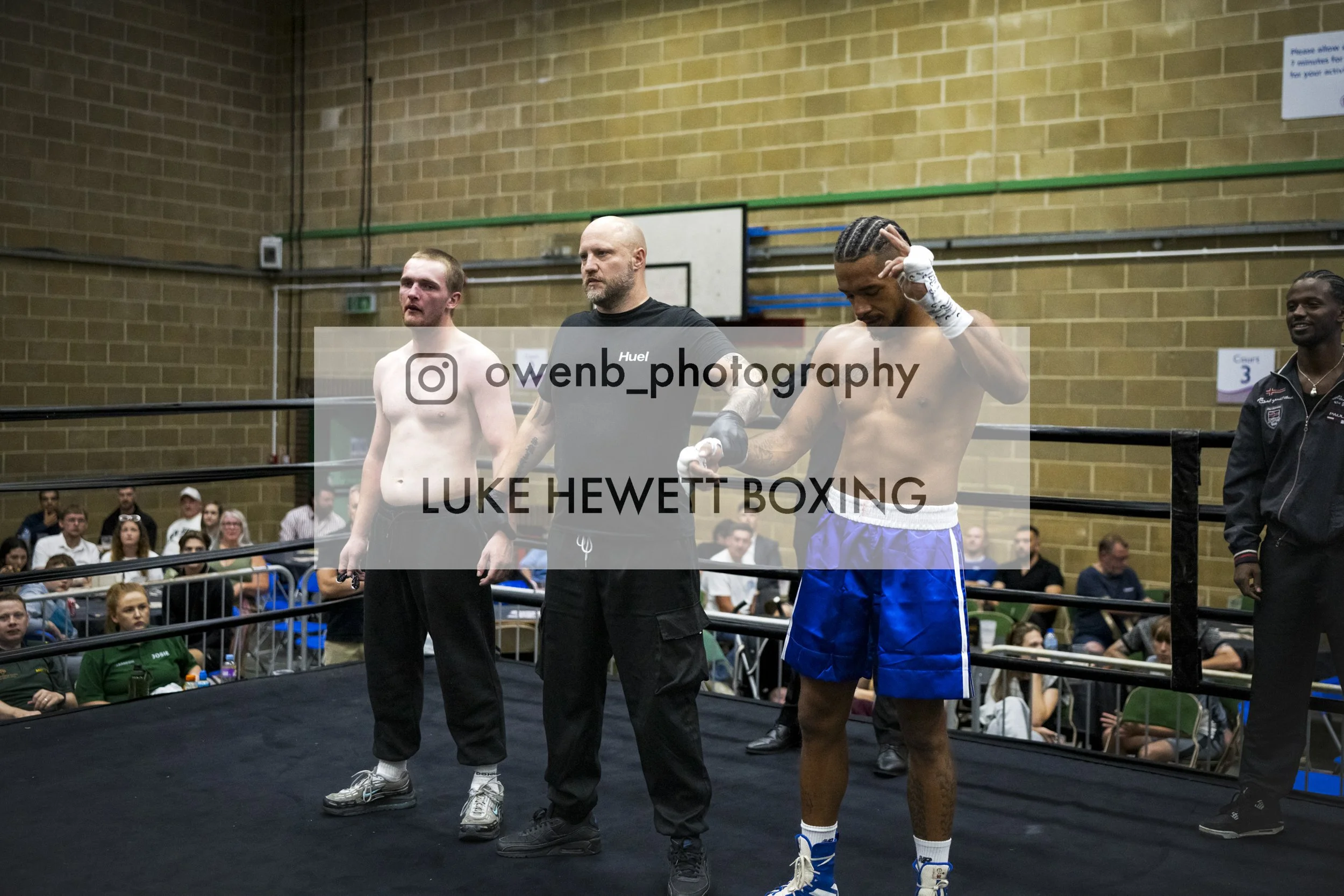 Boxing match scene inside a gymnasium with an audience. Two shirtless male boxers, one with a bandaged hand, are accompanied by a referee in black. The audience watches from seats, and the setting features a brick wall and sporting equipment.