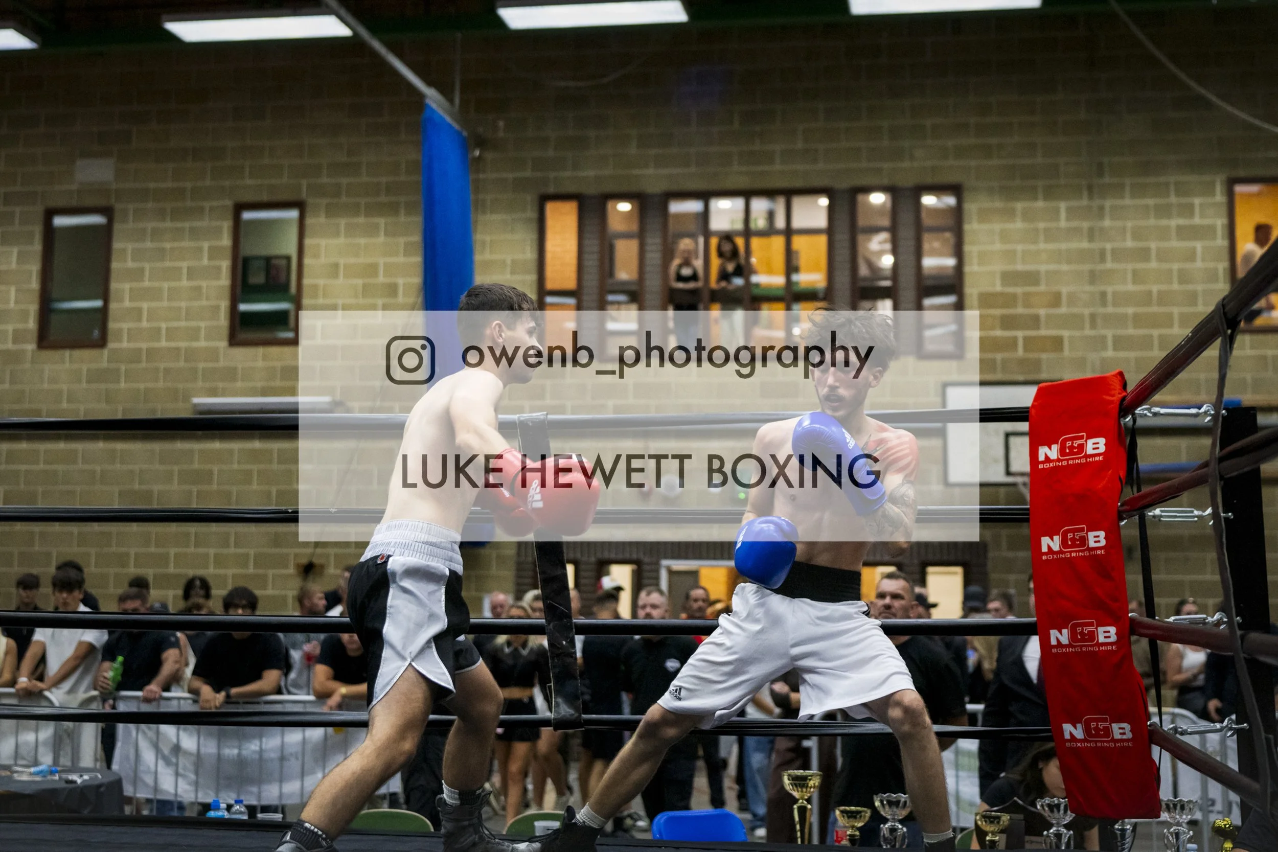 Two male boxers sparring in a boxing ring with an audience watching, trophies on a table nearby, and a large window showing the room's interior.
