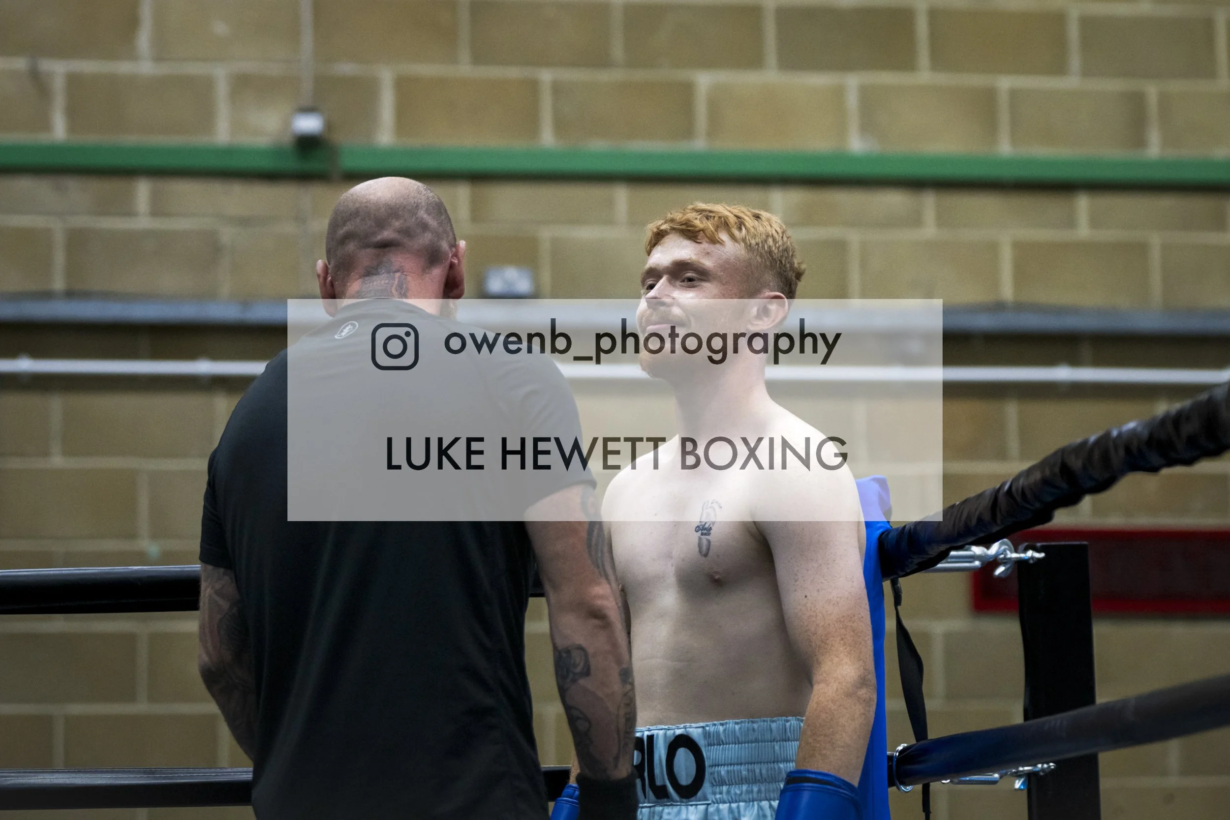 A young male boxer with red hair and tattoos on his arm in a boxing ring, talking to a coach or trainer.