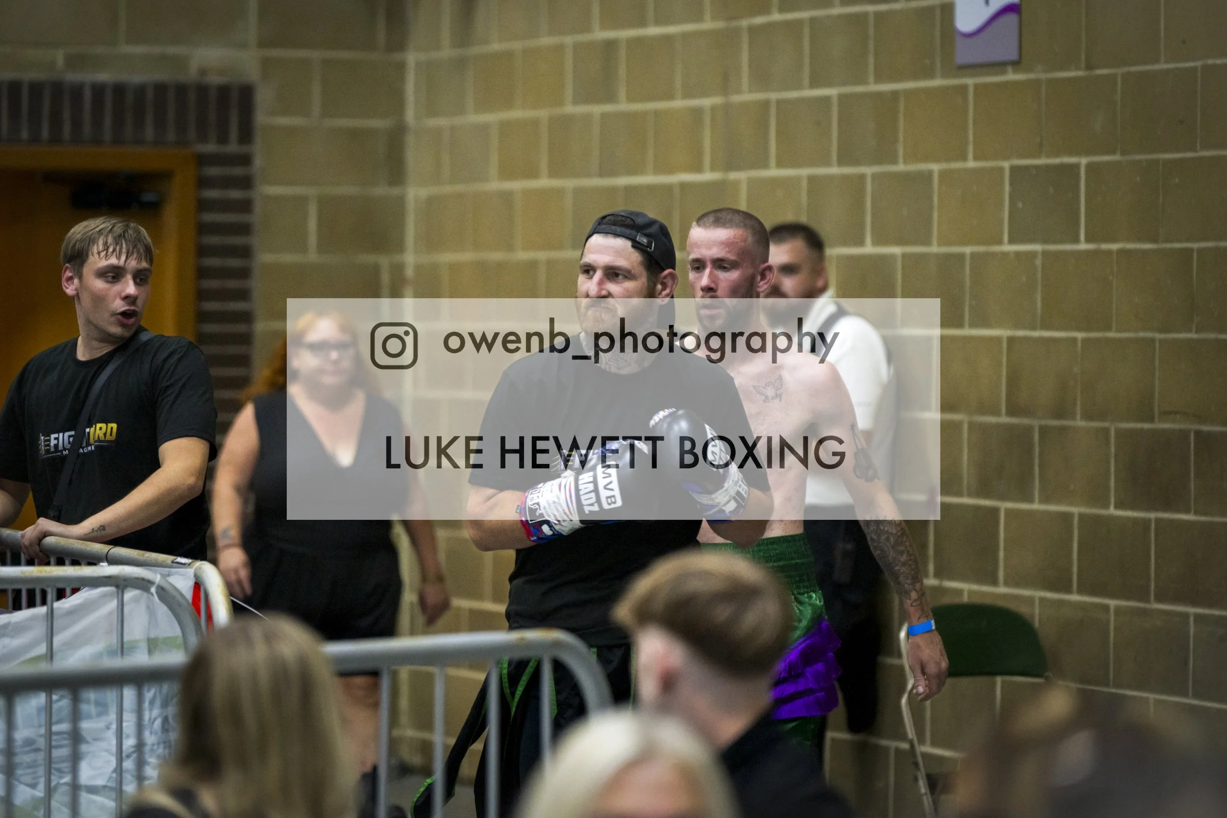 Boxer Luke Hewlett standing with others in a gym or event space, wearing gloves and green shorts, with a tattoo on his chest and a serious expression.