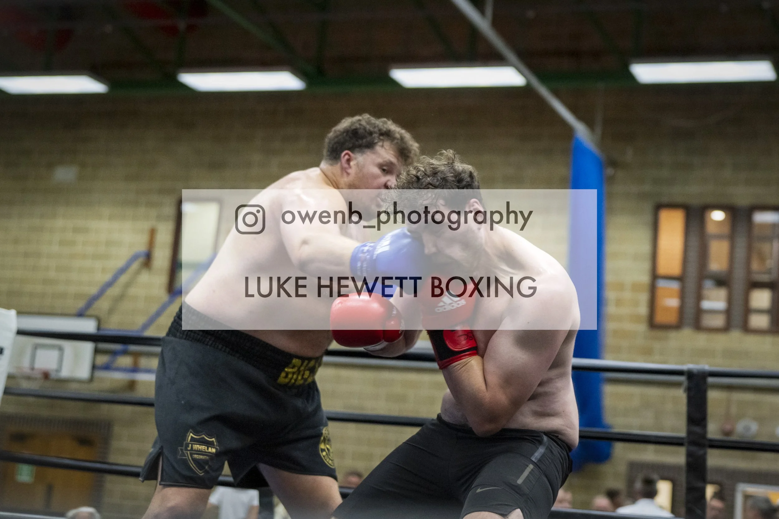 Two men engaged in a boxing match inside a gym. The man on the left, with curly hair, is wearing black shorts with yellow lettering and red boxing gloves. He appears to be attacking or applying a clinch to the other man, who has dark hair and is wear