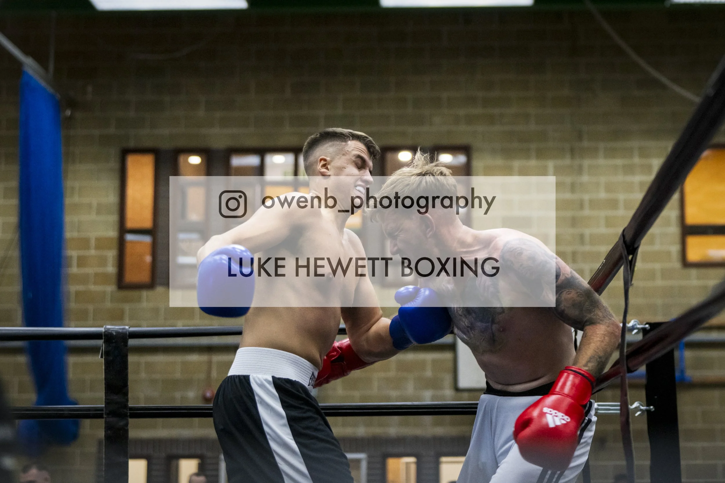 Two shirtless male boxers wearing gloves sparring in a boxing ring with a brick wall background.