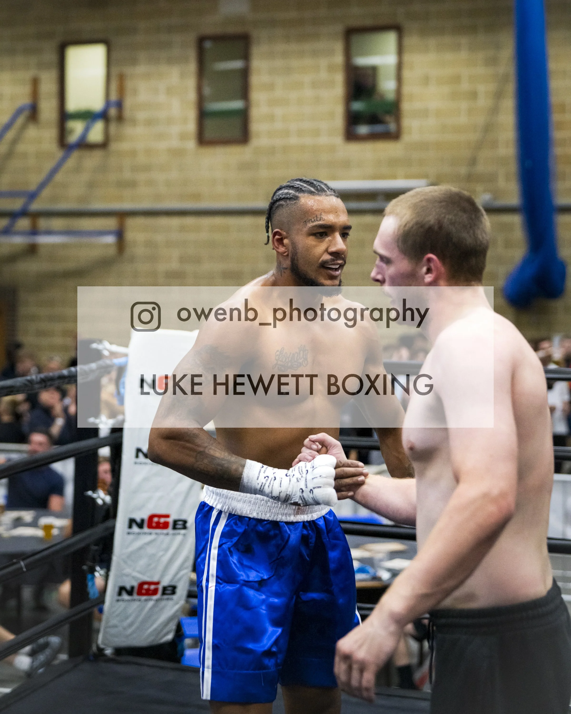 Two male boxers shaking hands inside a boxing ring, the boxer on the left has long braided hair and tattoos on his face and chest, and is wearing blue shorts. The boxer on the right has short hair and is shirtless. Several spectators are visible in t