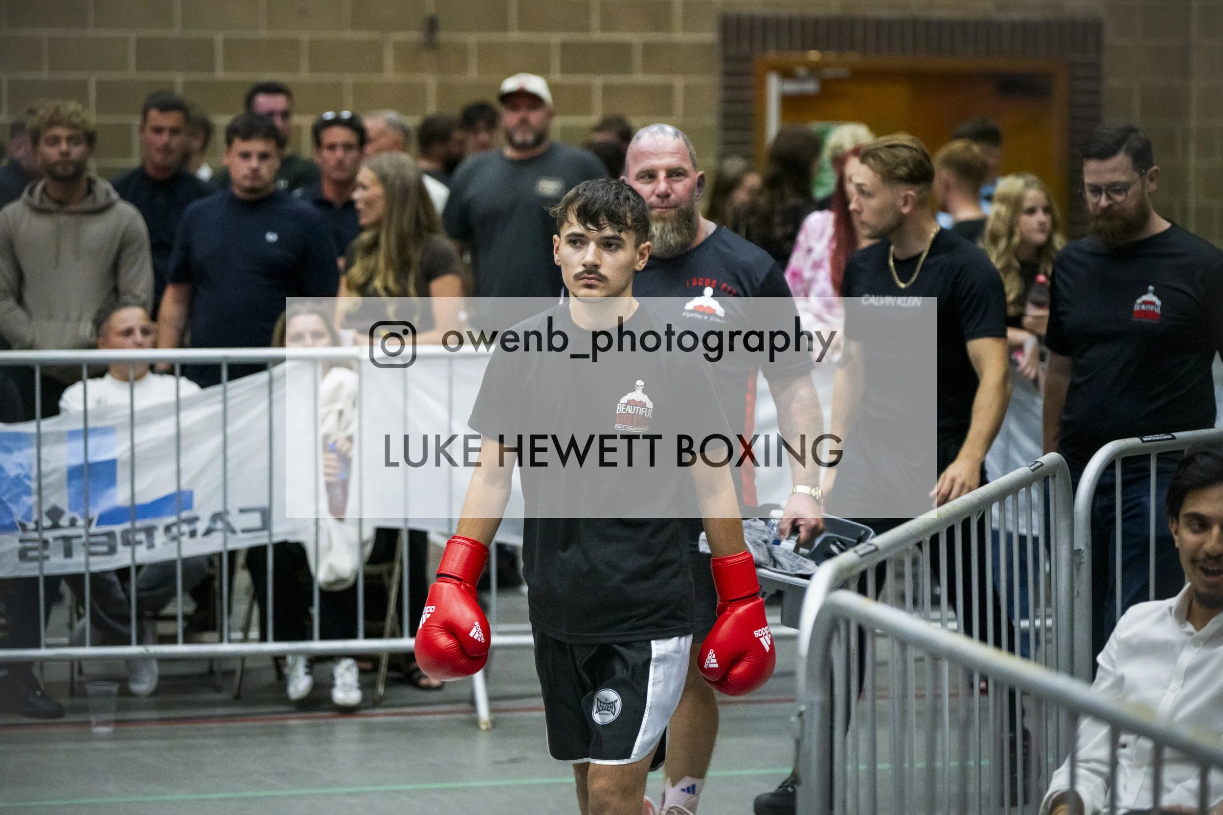 A young male boxer wearing red gloves and black and white shorts walking through a crowd with spectators behind metal barriers, at a boxing event.