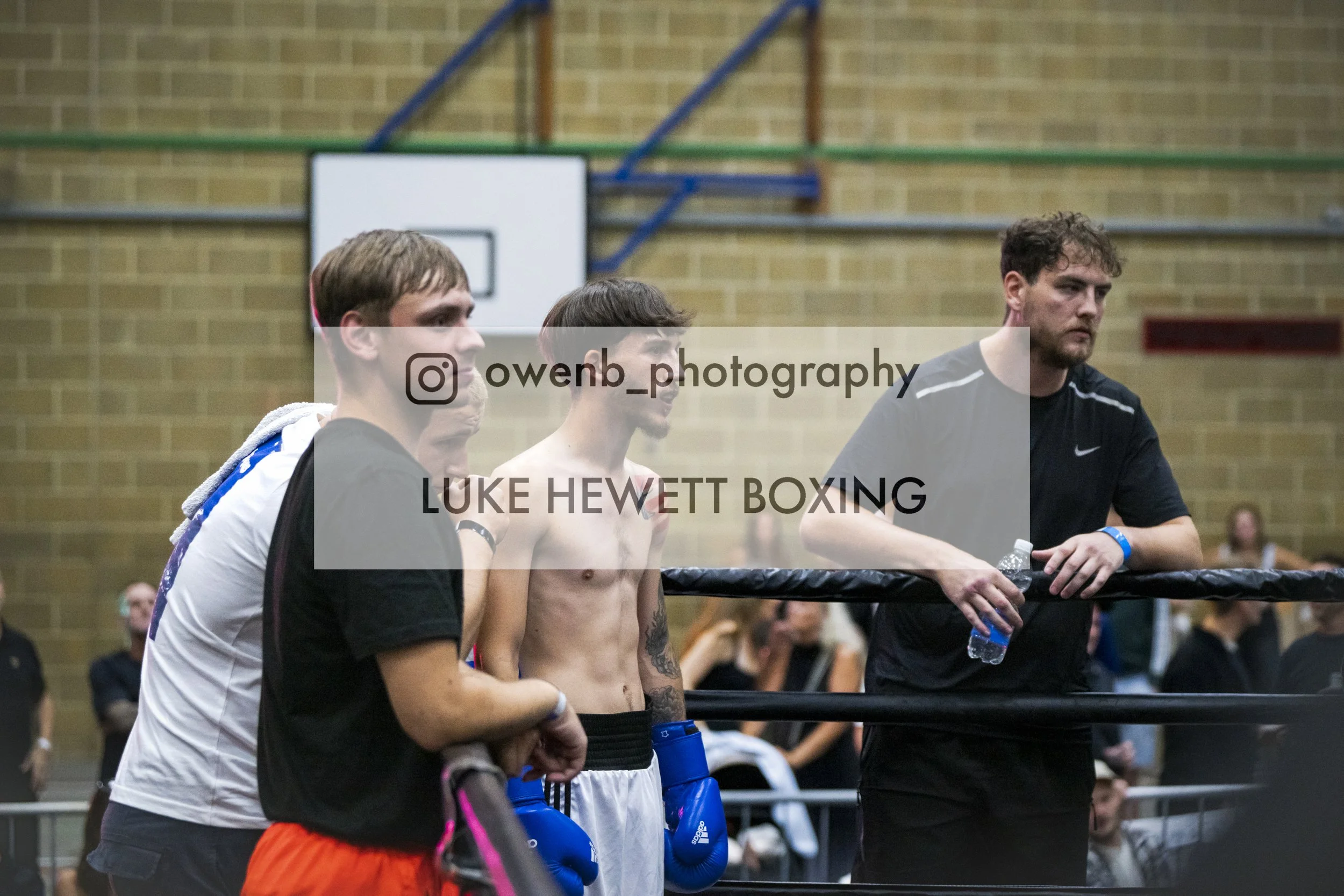 A group of boxers and a coach inside a gymnasium before a match, standing in a row with their hands behind their backs, watching a match with a boxing ring in the background.
