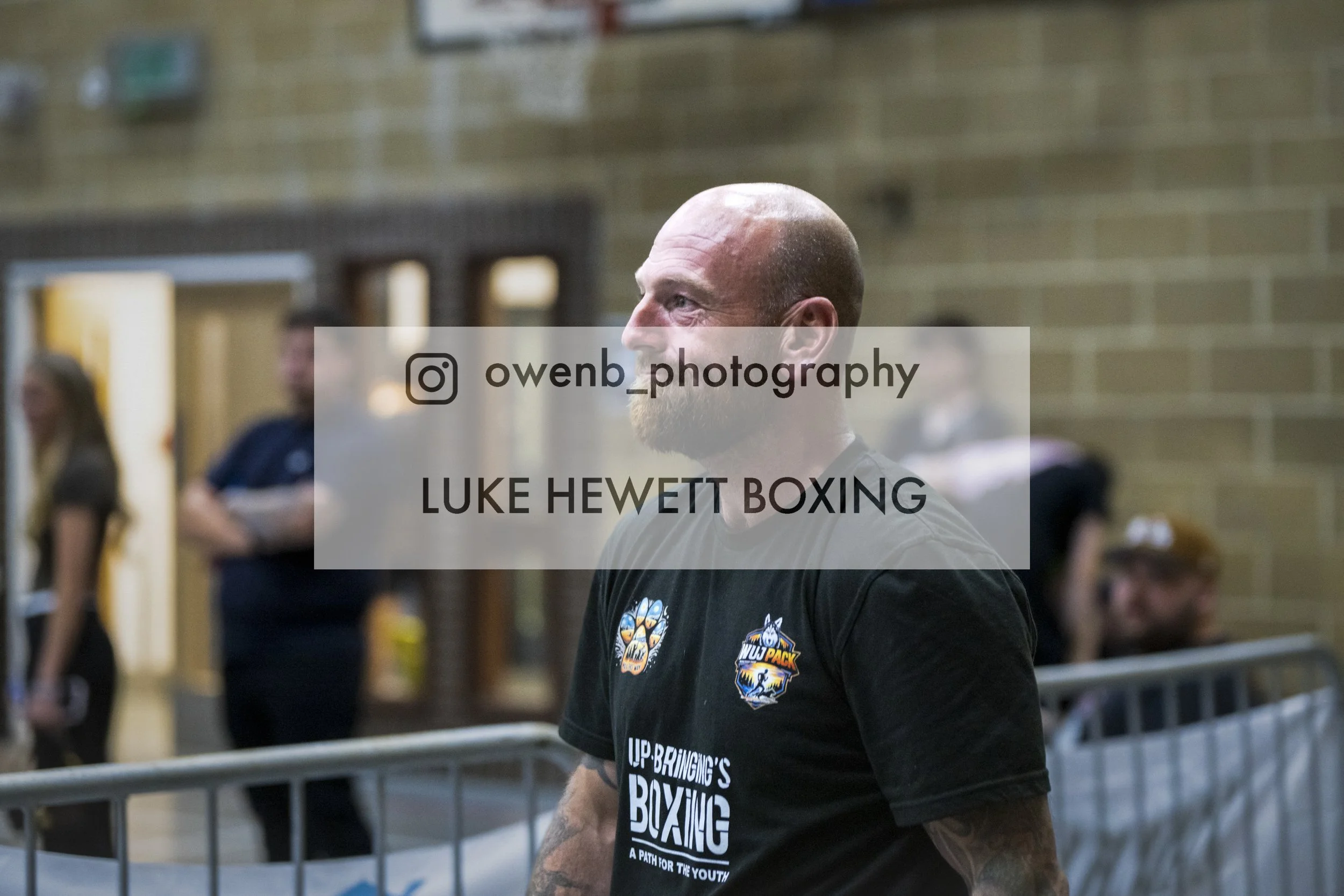 A man with a shaved head and beard, wearing a black T-shirt with a boxing logo, stands in a gymnasium with a brick wall, metal railings, and blurry people in the background.