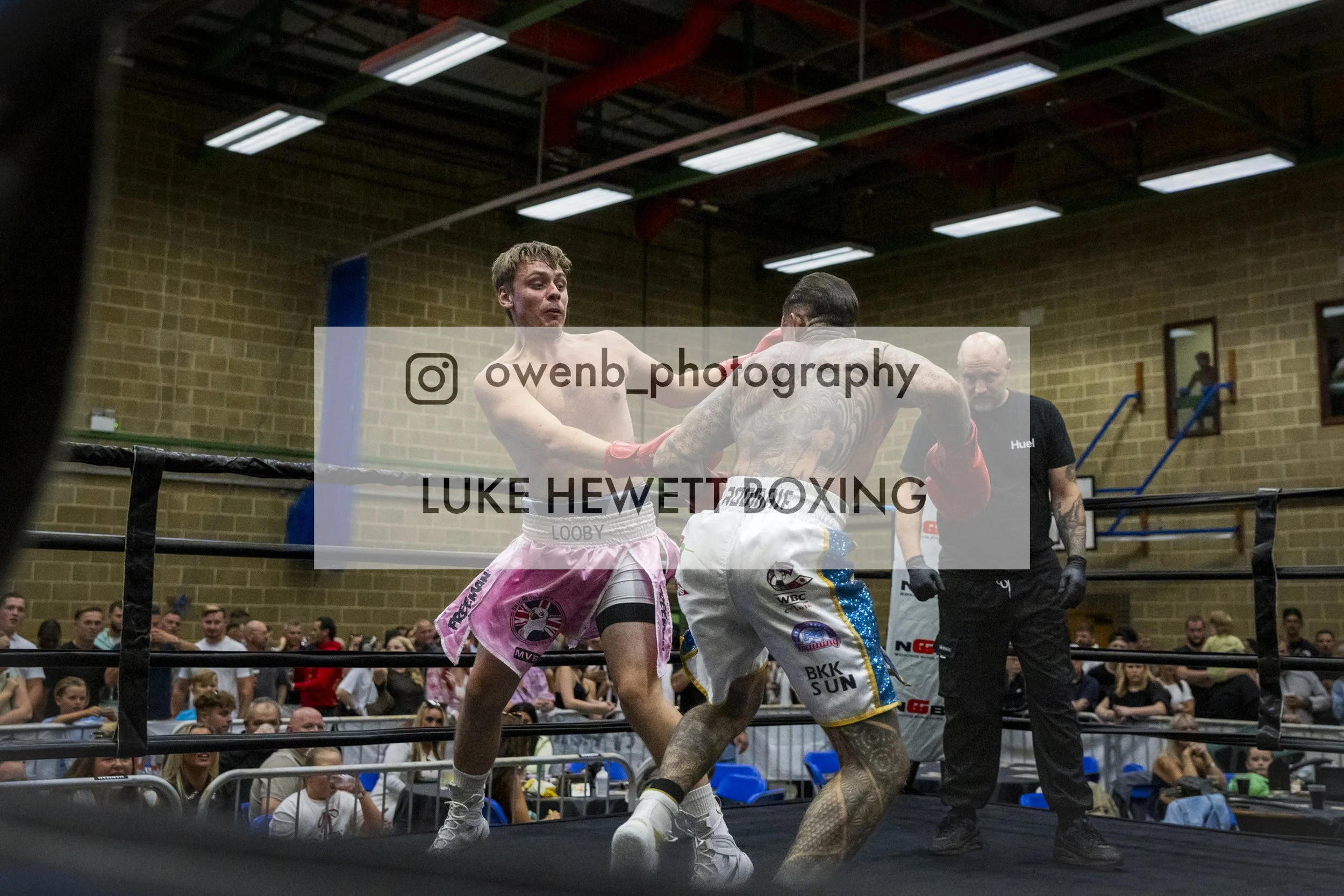 Two male fighters in a boxing ring with a crowd watching. One fighter is wearing pink shorts and the other white shorts with colorful patterns, and they are in the middle of a match. A referee is present behind them.
