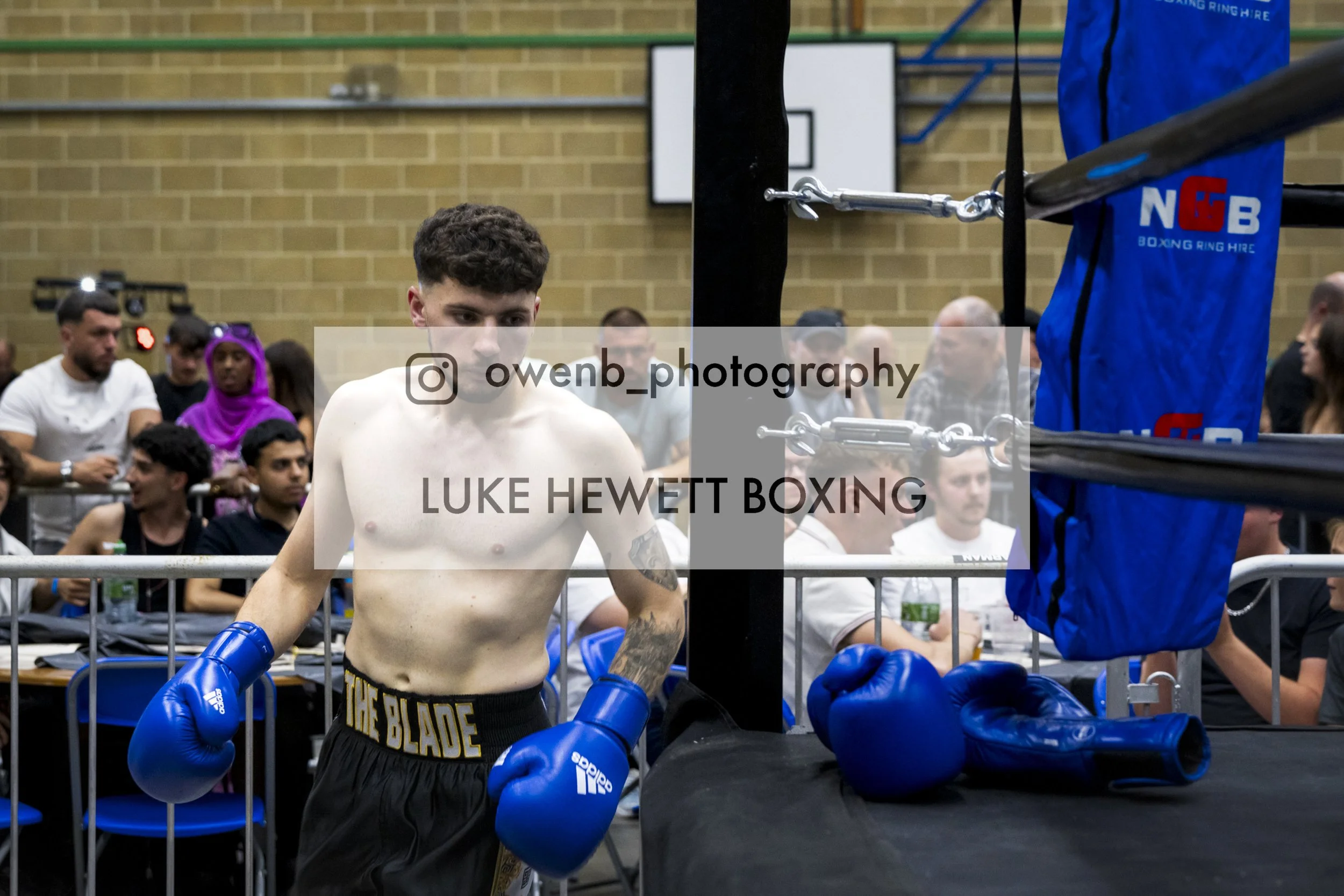 A shirtless male boxer with blue gloves inside a boxing ring, surrounded by spectators and officials, during a boxing match or event.