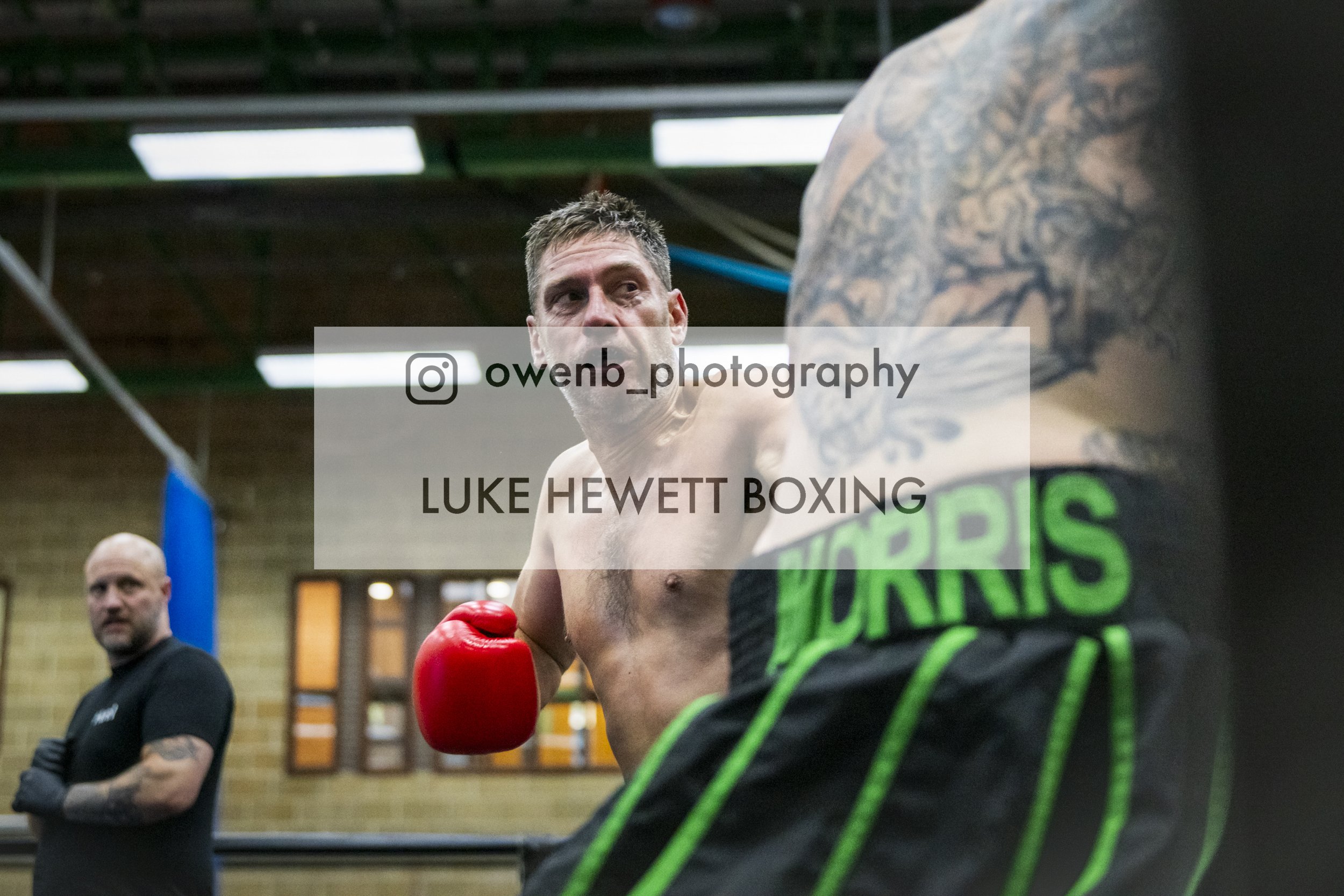 A boxer with tattoos on his upper body and red boxing gloves, looking towards an opponent in a boxing ring. Another man with a tattooed arm and dark shirt stands in the background. The setting appears to be a gym or training facility.