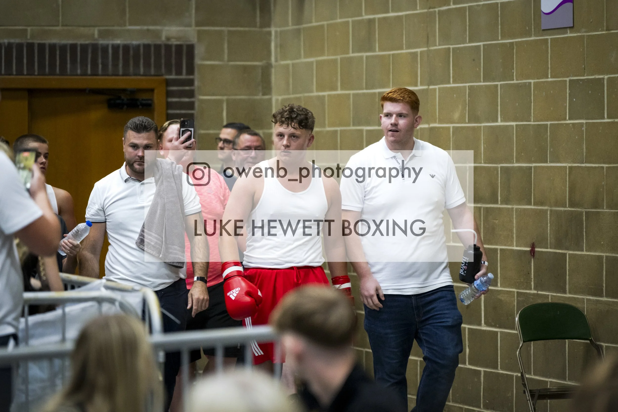Boxer in red shorts and gloves walking through an indoor room, surrounded by spectators and staff, with a man in white shirt holding water bottles and a towel.