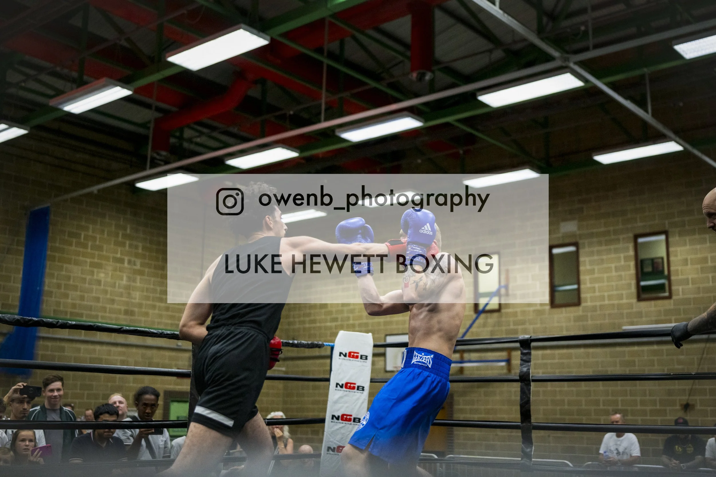 Two boxers in a boxing ring engaged in a match, with one delivering a punch to the other. The scene is indoors, with a brick wall and spectators watching.