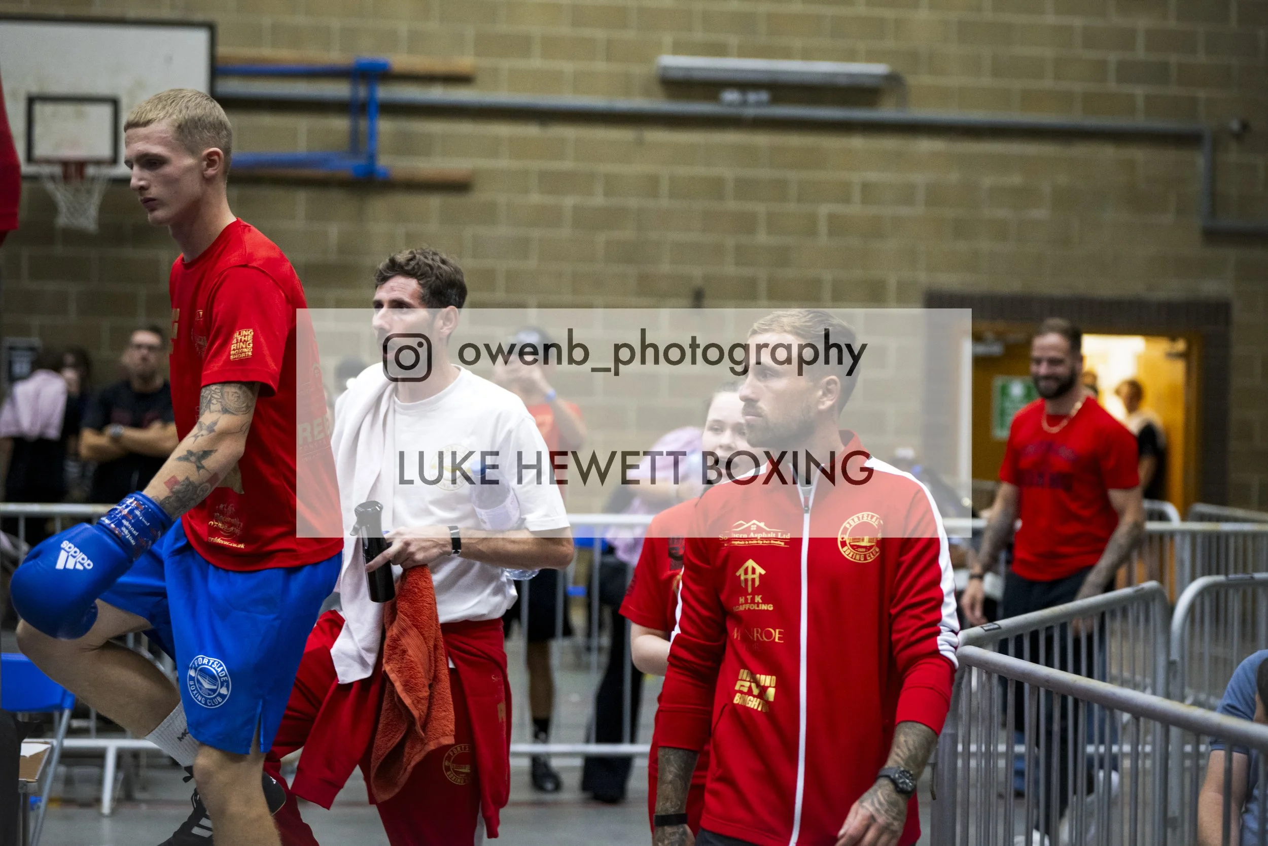 Boxer in red shorts and shirt, with tattoos on his arm, preparing to fight in a boxing ring with several people around, including spectators and officials.