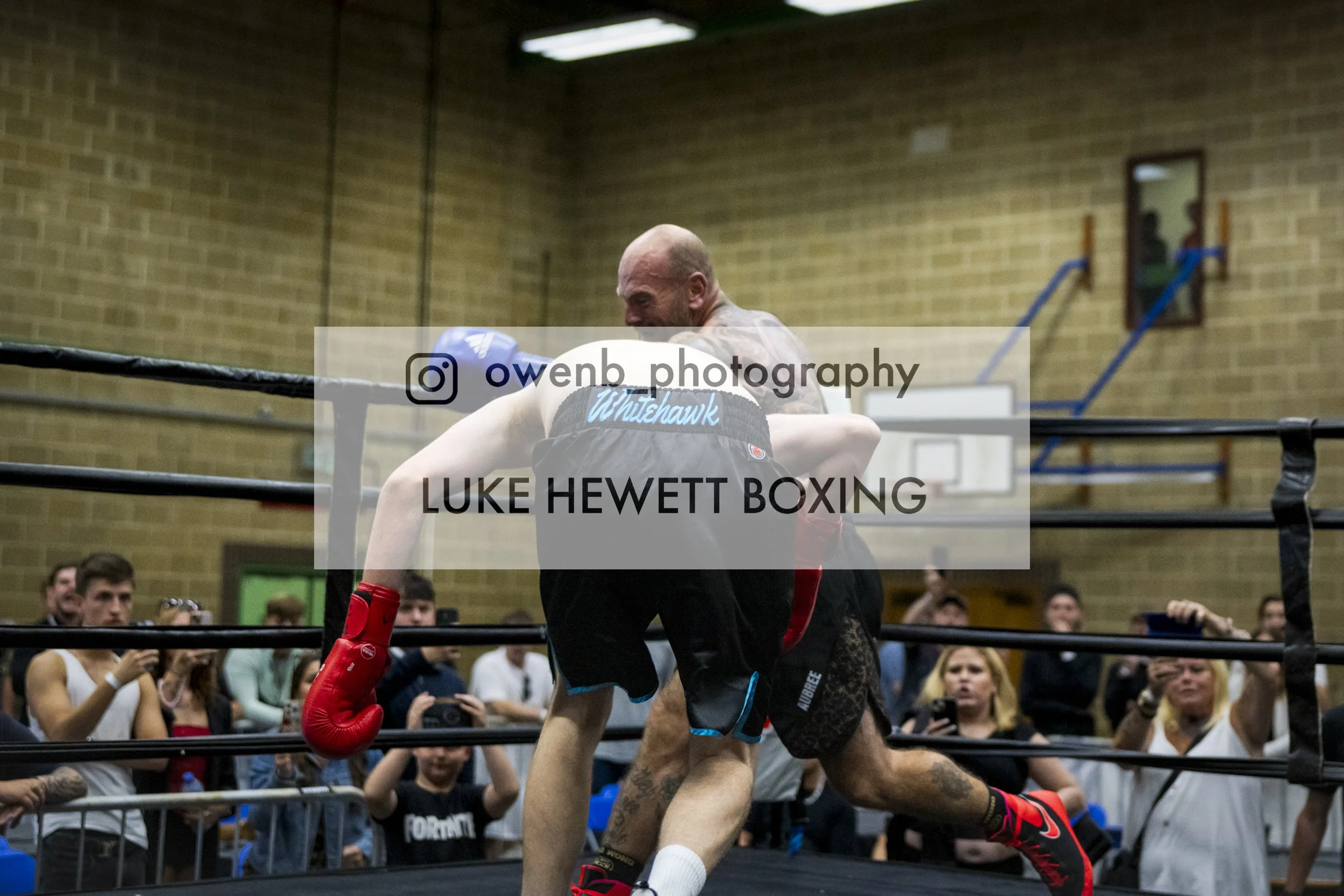 Boxer in black shorts and red gloves in a boxing ring, with a crowd watching against a brick wall in the background.