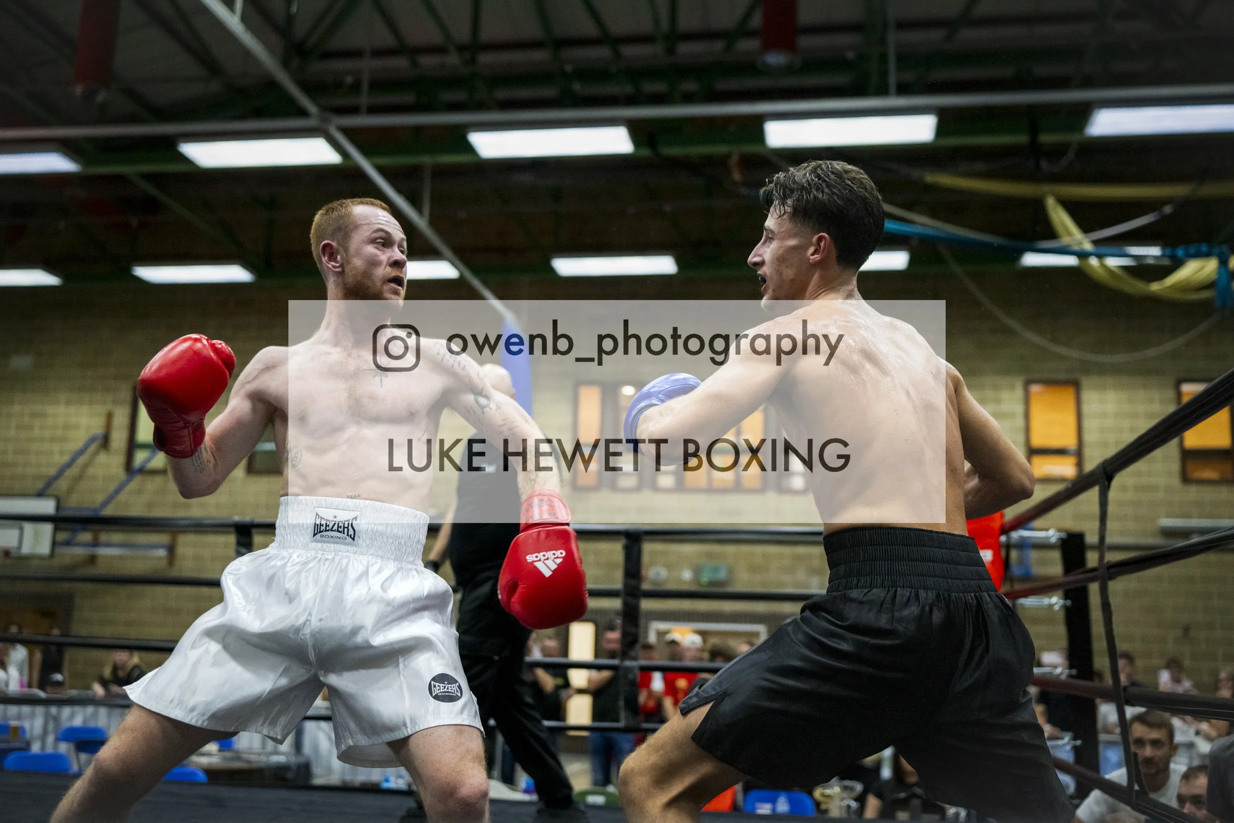 Two shirtless male boxers in gloves face each other in a boxing ring during a match, with one in white trunks and the other in black trunks, in an indoor gym.