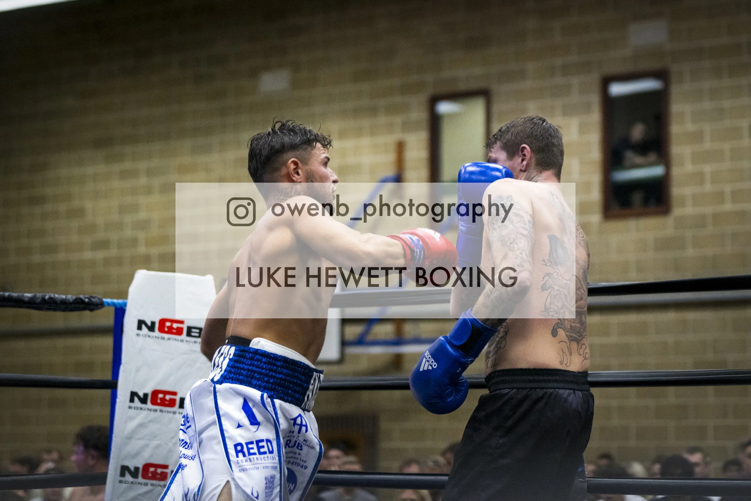 Two boxers in a boxing match. One is throwing a punch while the other is blocking. The boxer on the left is shirtless with blue boxing gloves and shorts, the boxer on the right has tattoos and is wearing blue gloves. The scene is inside a boxing gym 