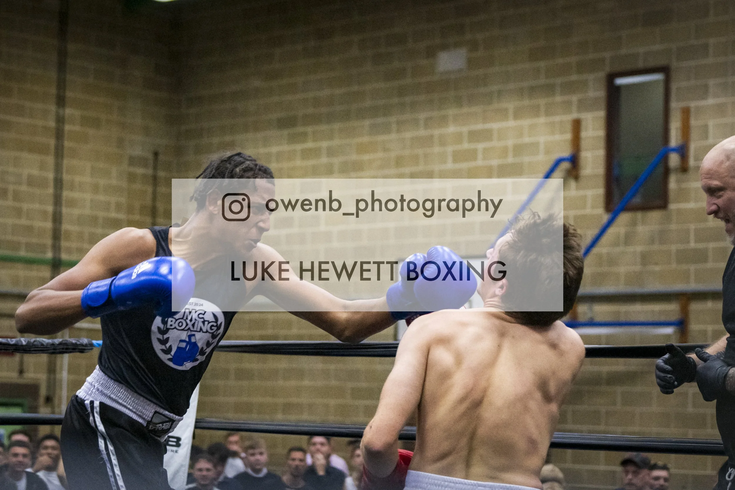 Two male boxers exchanging punches in a boxing match, one wearing blue gloves and the other shirtless, with an audience watching in the background.