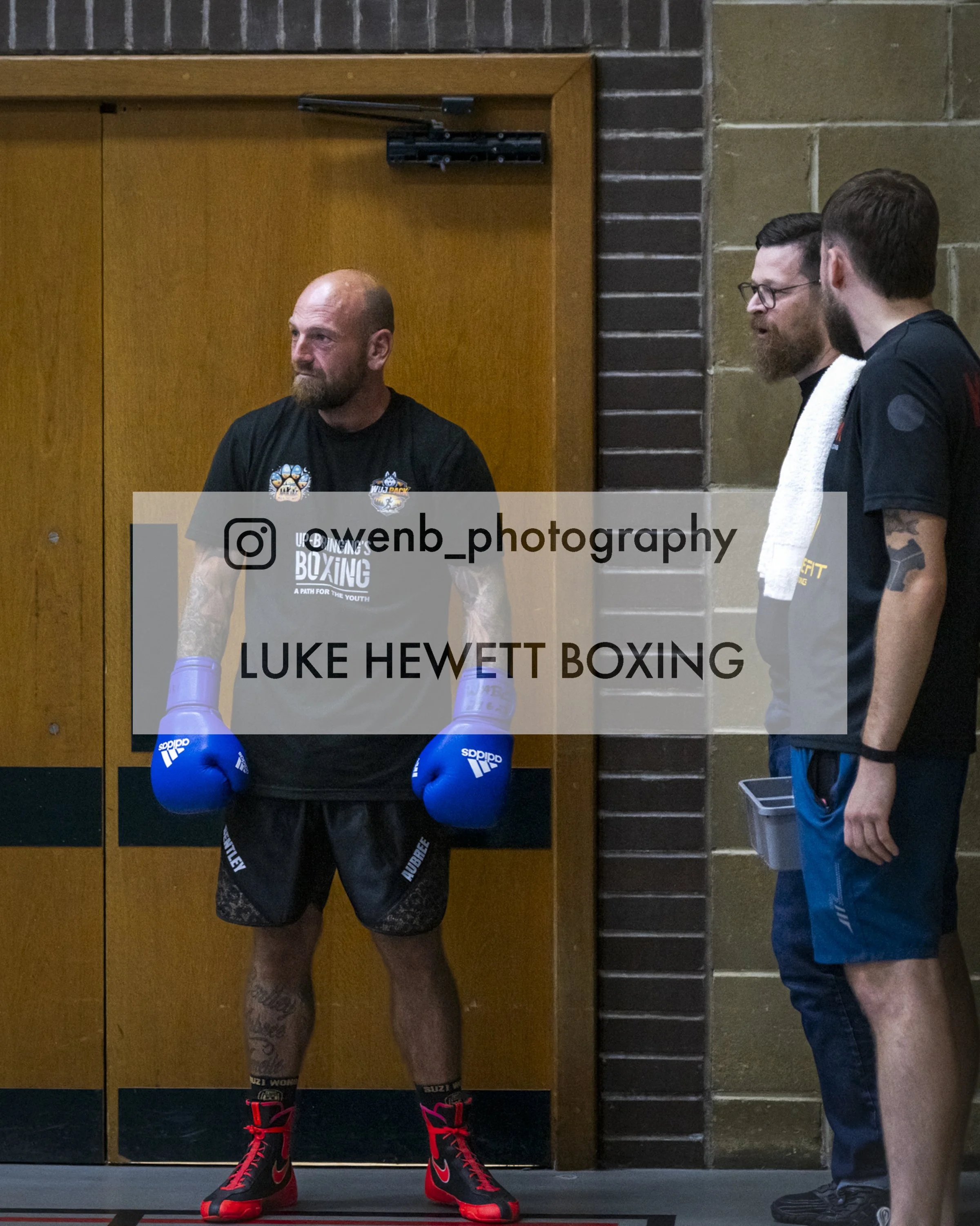 Boxer Luke Hewett wearing black boxing shoes with red laces, black shorts, and boxing gloves, standing in a gym with two men talking nearby.