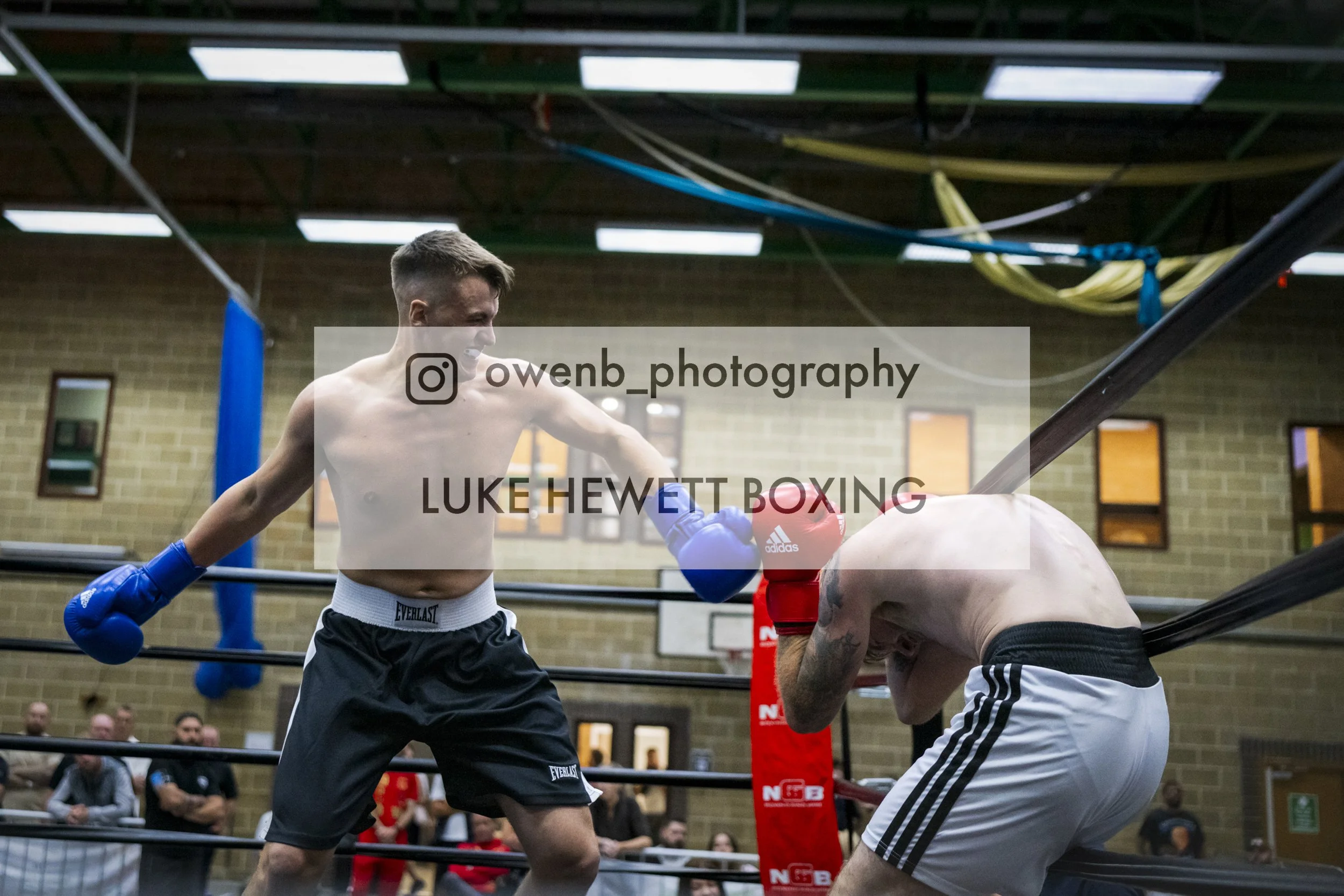 Two shirtless male boxers with gloves fighting in a boxing ring, one delivering a punch to the other, with spectators watching in the background.