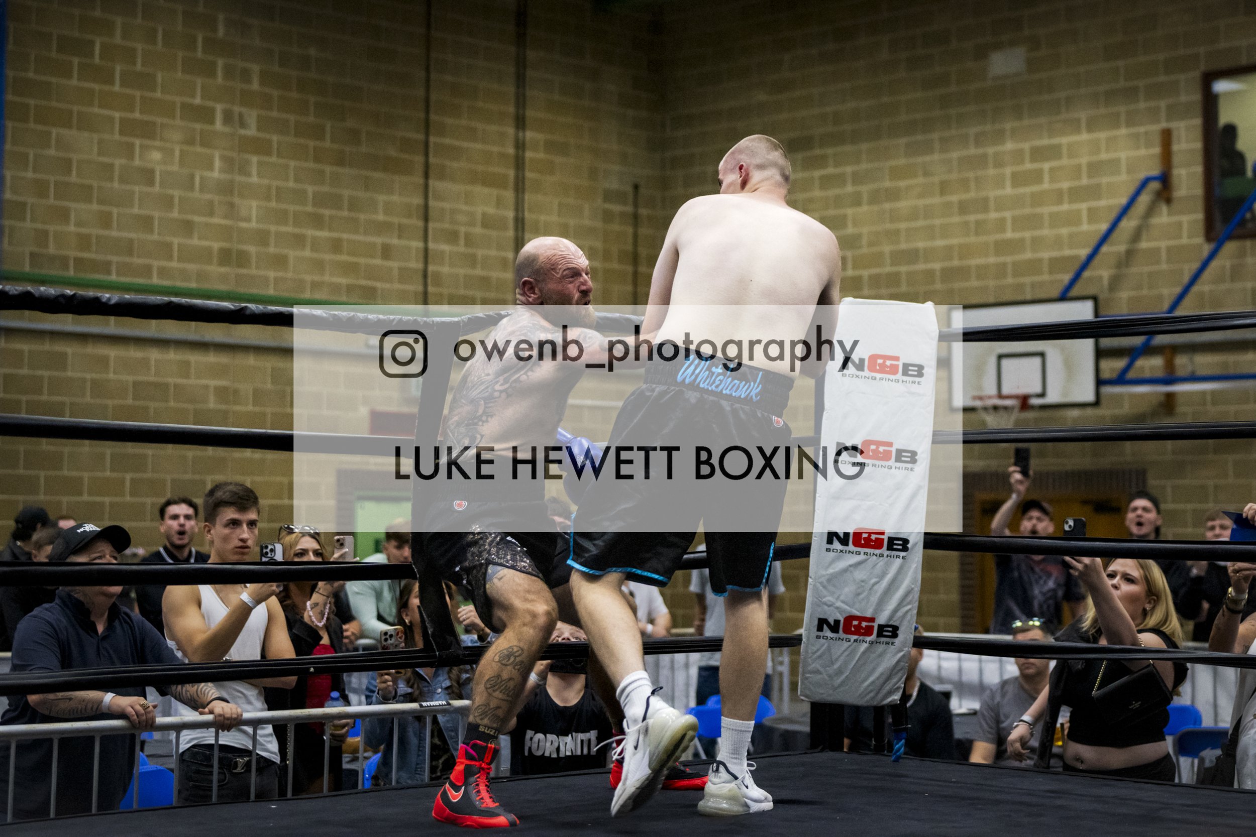 Boxers training in a ring with an audience watching, surrounded by a brick wall, at a boxing event.