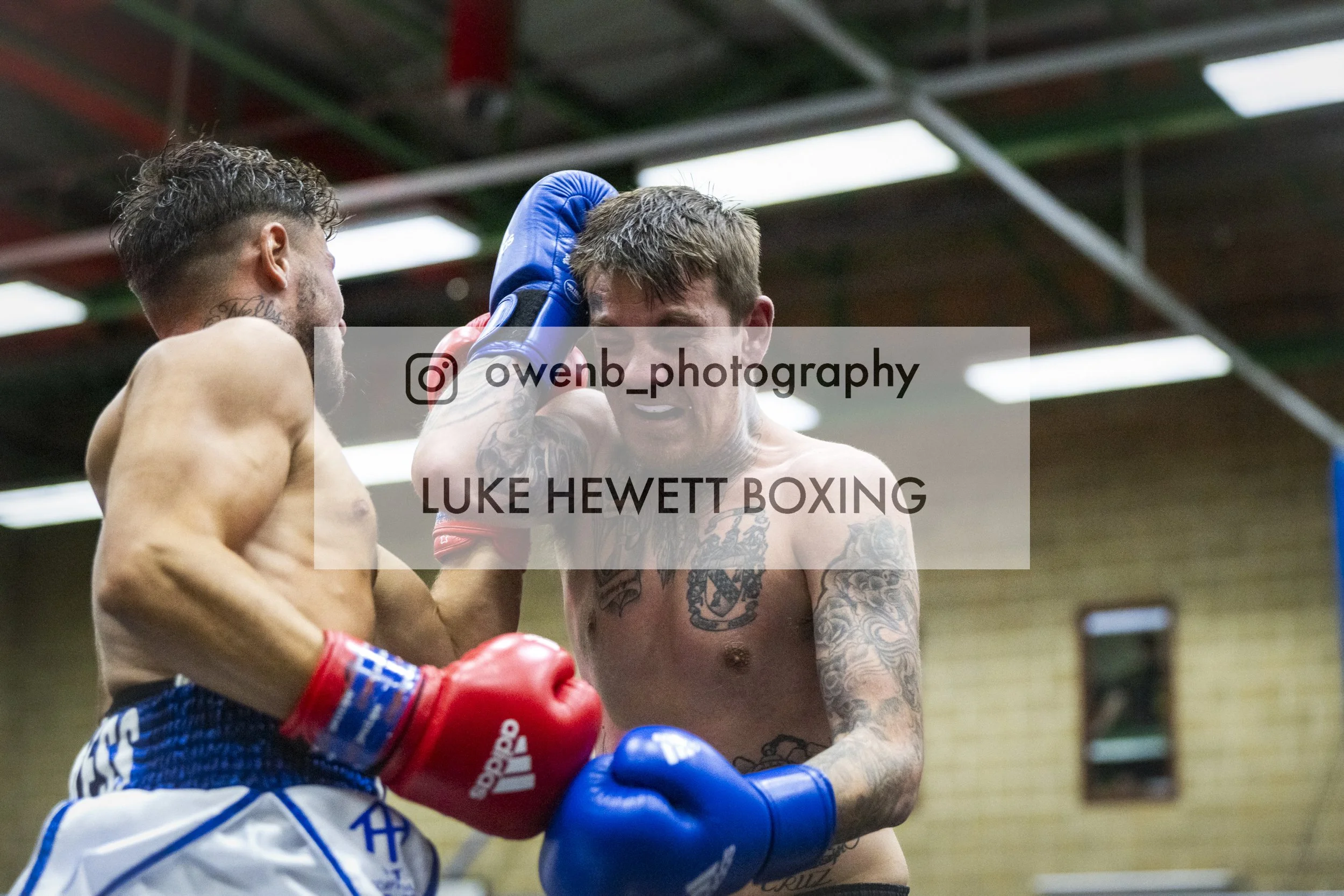 Two shirtless men with boxing gloves engaged in a boxing match inside a gym. One man is holding the other near the head with his glove. The man being held has tattoos on his chest and arm. The background shows a gym ceiling with lights.