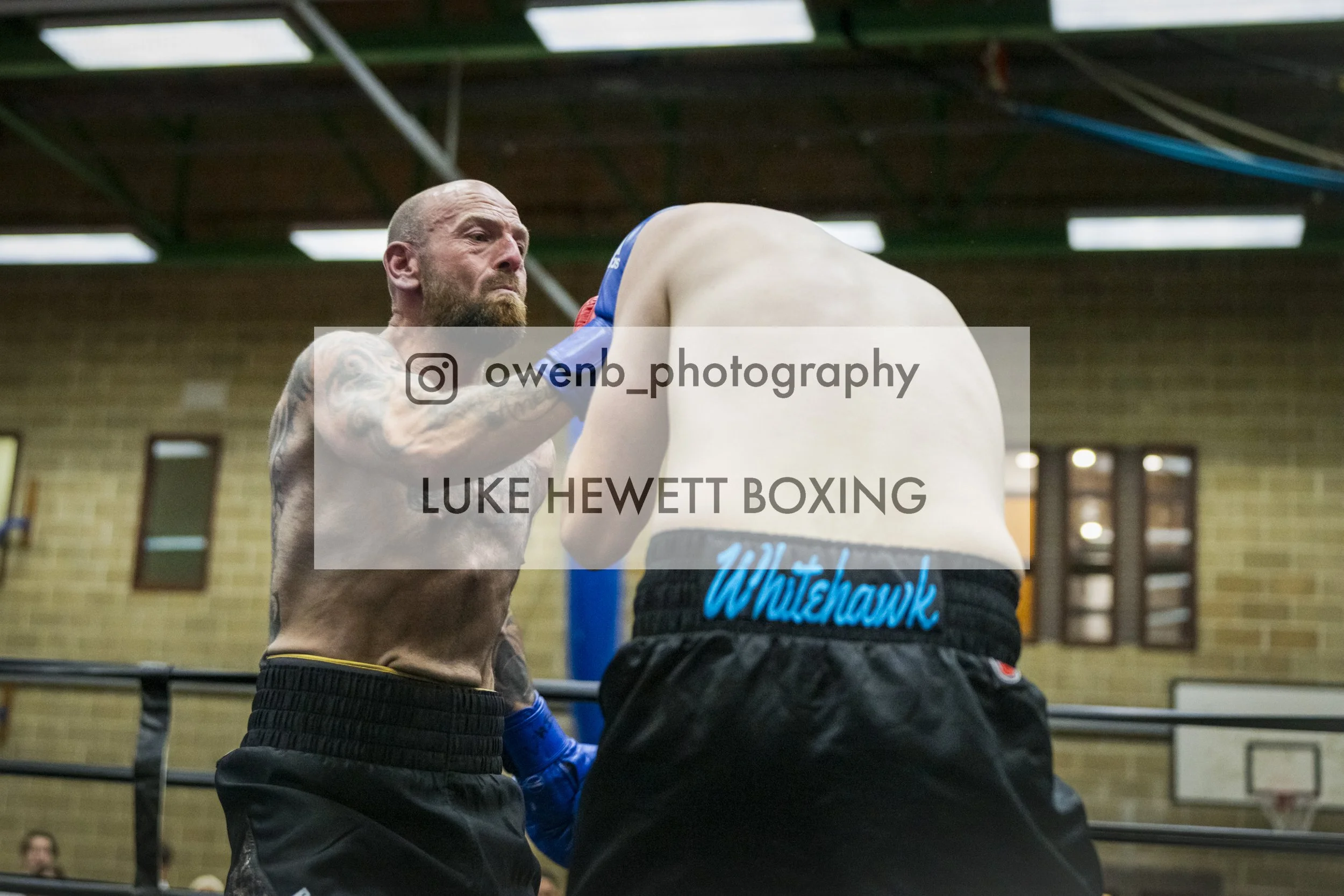 Boxer with tattoos on arm in boxing ring, facing another fighter with 'Whitehawk' on waistband.