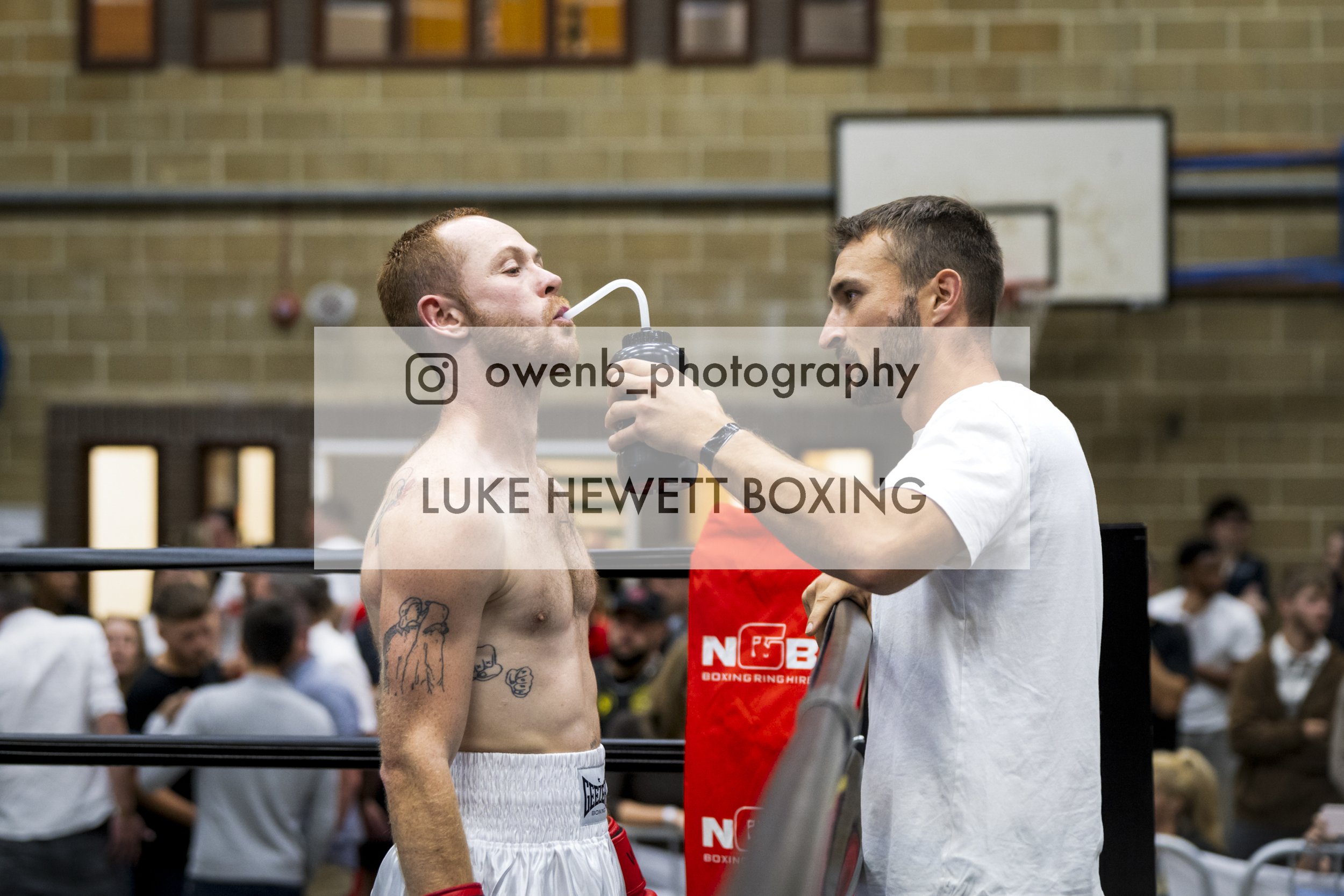 A shirtless boxer with tattoos on his left chest and arm drinks water through a straw held by another man in a white shirt. They are inside a gym with a brick wall and a basketball hoop in the background, and a crowd of people watching.